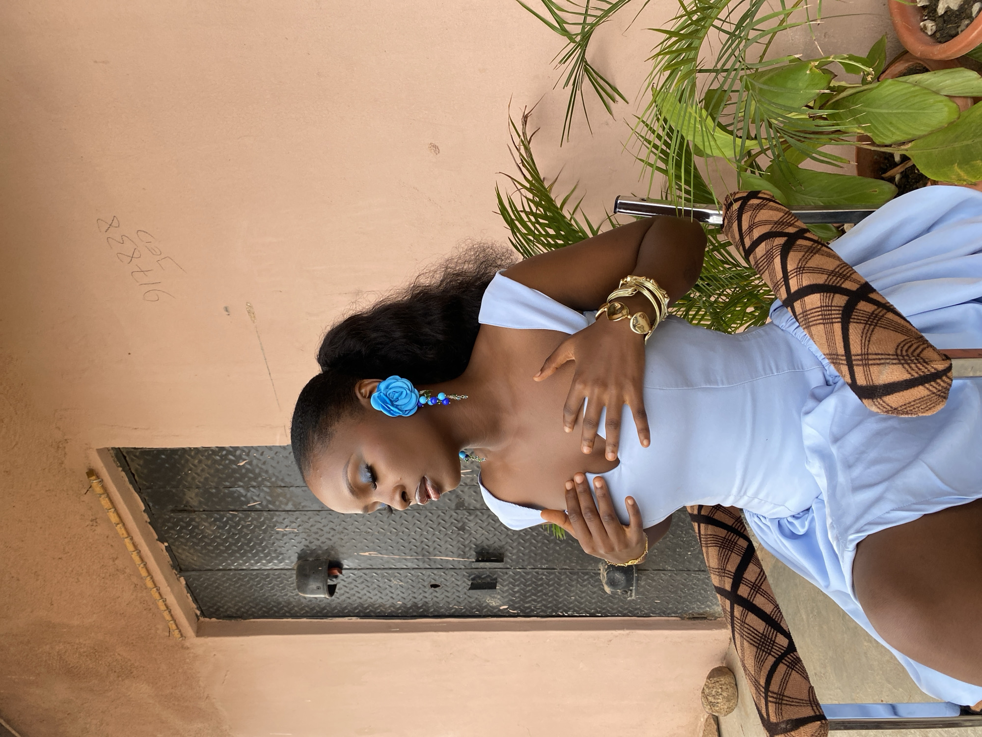 A dark-skinned woman with dark hair in a ponytail, wearing a light blue dress, sits in a chair in front of a textured pink wall. She has large blue flower-shaped earrings and gold bracelets on both wrists. Her eyes are closed, and her hands are placed on her chest. To her left is a metal door with diamond plate texture. In front of the wall, to the right of the woman, are two potted plants. The wall has some graffiti that appears to be a phone number and the word "JED". The ground is a dirt path. The overall scene appears to be outdoors in a somewhat urban environment, possibly in Jos, Nigeria, given the graffiti.