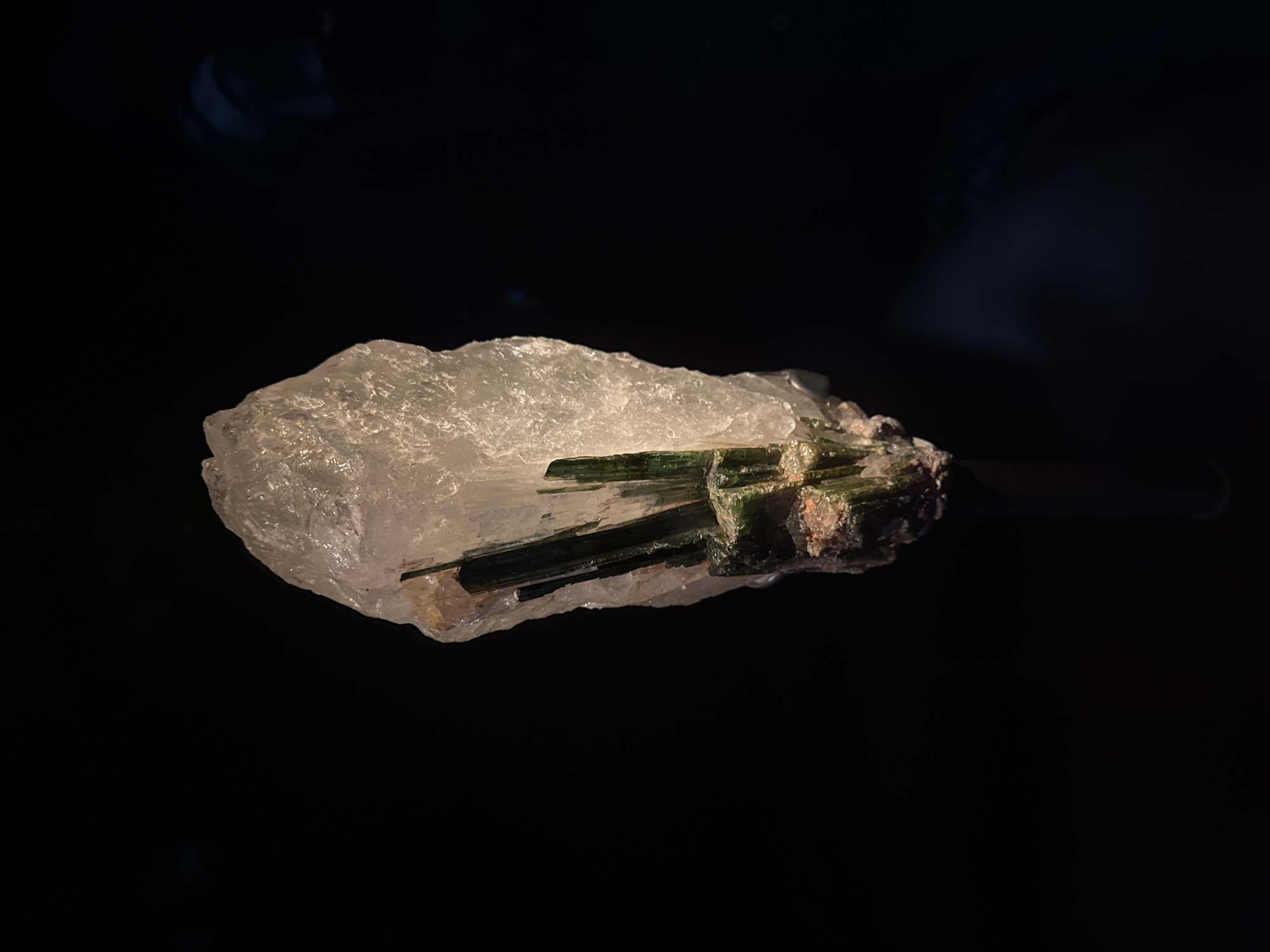 A dark, studio-lit shot features a specimen of green tourmaline crystals embedded within a rough, translucent quartz matrix. The specimen is centrally placed, with the dark background providing a stark contrast that emphasizes the texture and color of the mineral. No people or specific London landmarks are visible.