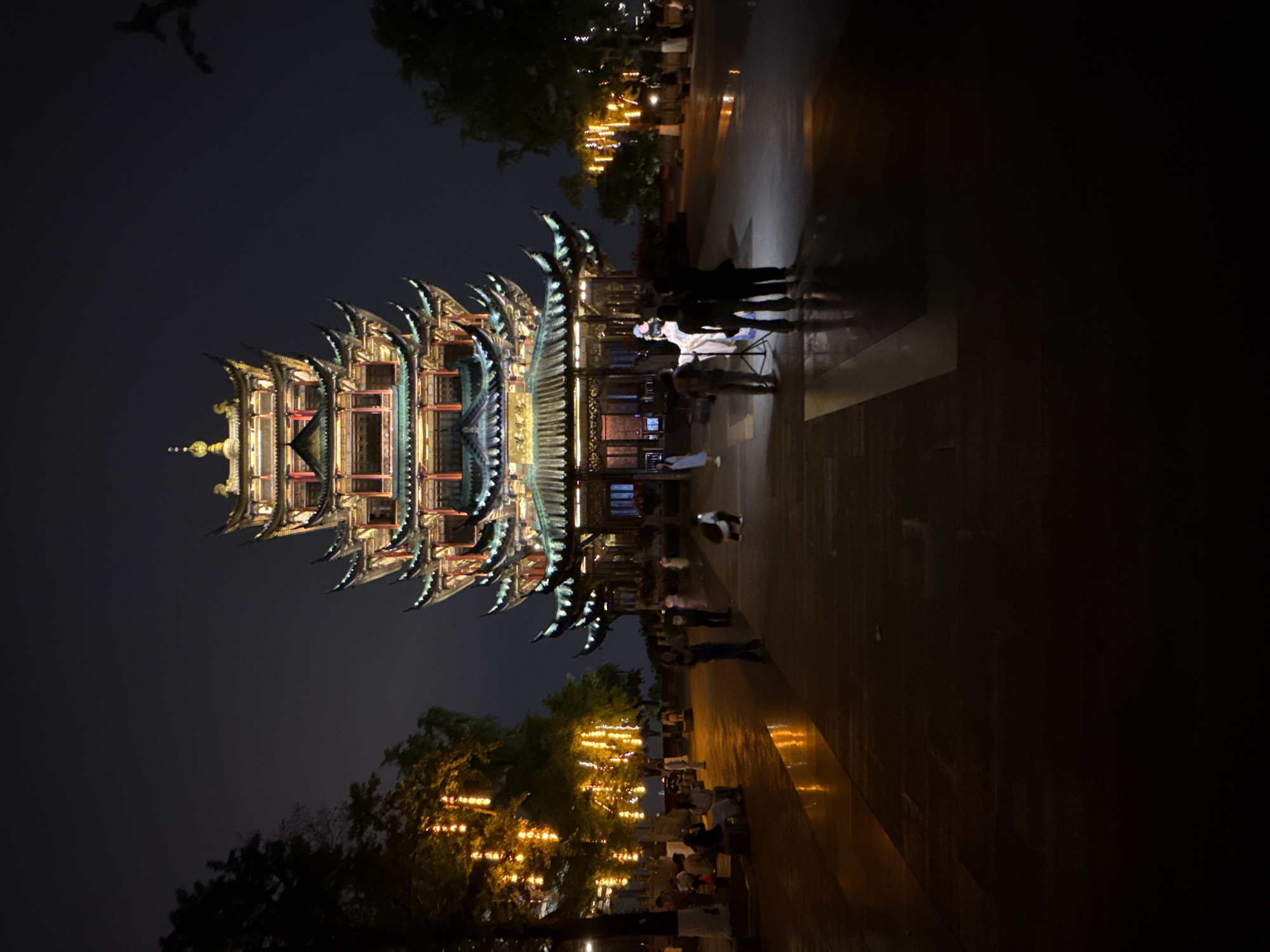 The image depicts a tall, ornate pagoda illuminated at night in Liangjiang, China. The multi-tiered structure features intricate architectural details, and rows of people are present, some standing near the pagoda and others gathered beneath trees adorned with decorative lights. The ground appears to be wet, reflecting the lights and creating a glistening effect. The surrounding environment consists of trees and other urban elements, all under a dark night sky.