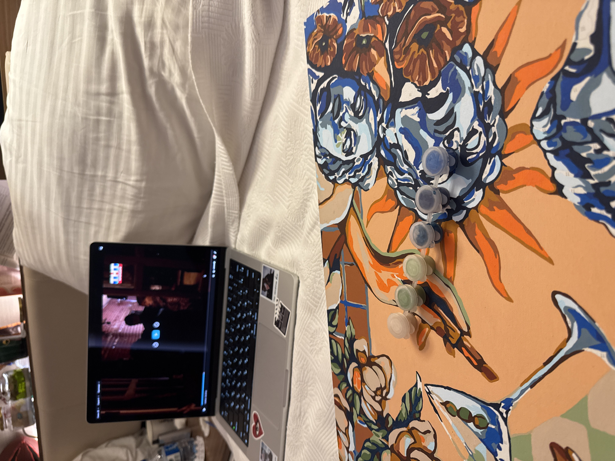 A laptop is open on a bed, displaying a video with a logo that reads "TNT". Next to the laptop is a paint-by-numbers canvas depicting a stylized image with floral elements and a face. Four small paint pots are placed on the canvas. The background consists of white bedding and a shelf with jars and bottles. There are no people in the immediate scene. The location is not specified as Aksu, Turkey, nor is there any information to infer this location.