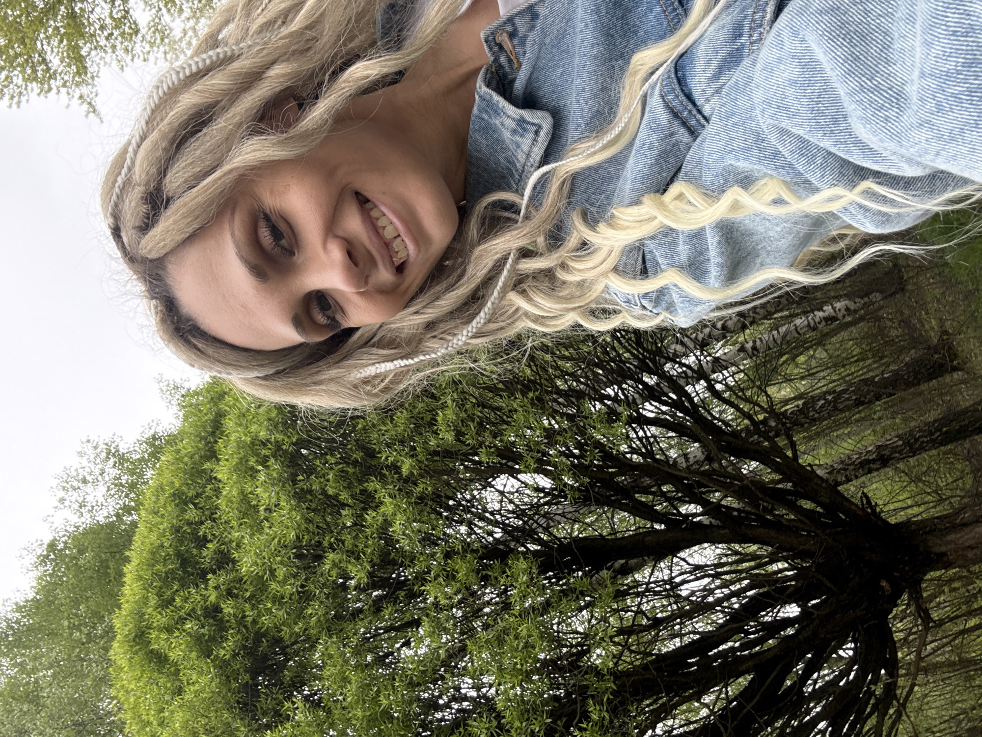 A woman with blonde dreadlocks and wearing a denim jacket smiles at the camera. The background includes green trees and a cloudy sky. This photo was likely taken outdoors in Dumino, Russia.