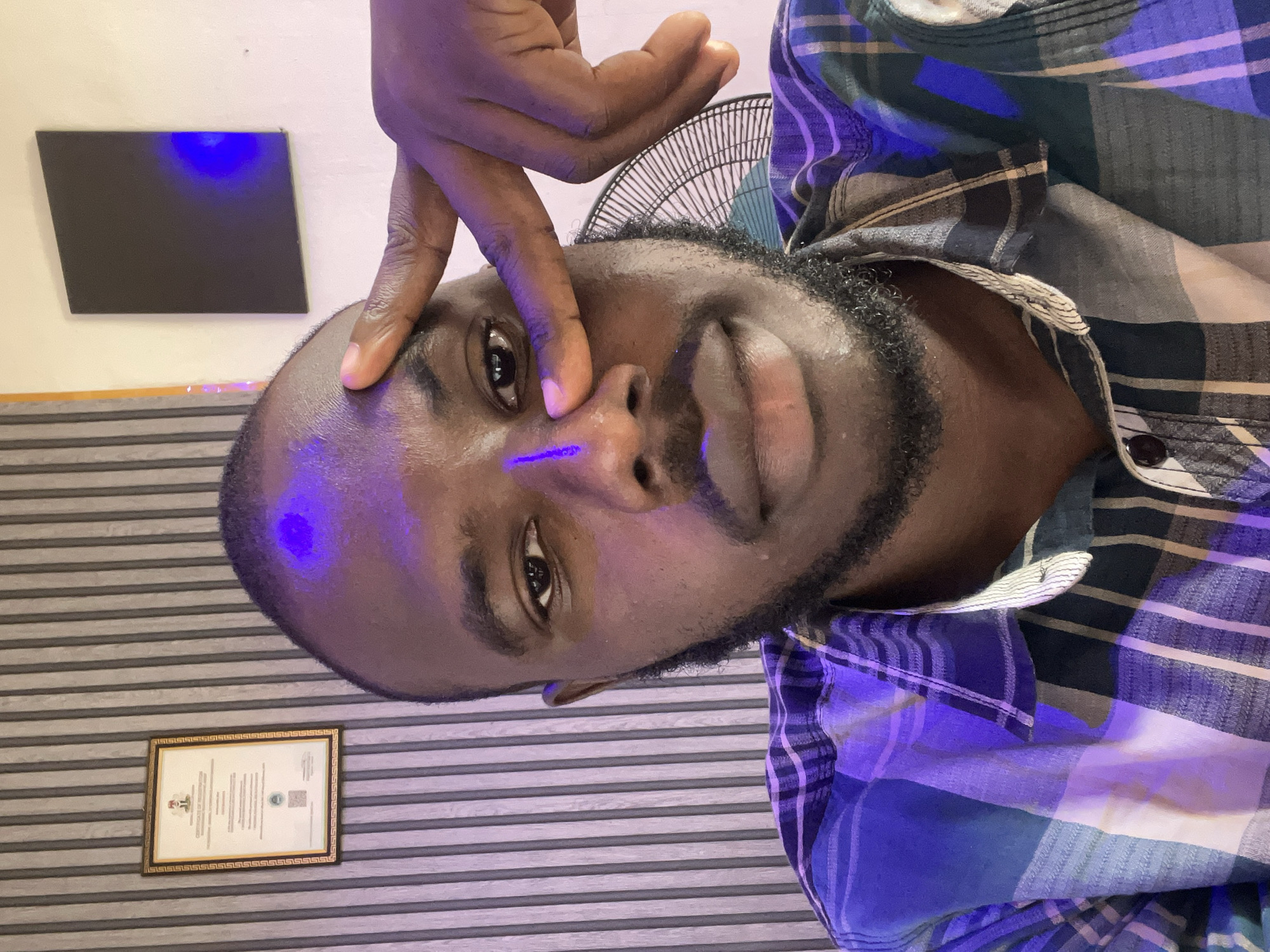The image is a close-up, selfie-style portrait of a dark-skinned man with a beard, positioned centrally in the frame. He is wearing a plaid shirt in shades of blue, white, and possibly green. His left hand is raised, with his index finger touching his forehead near his eye, and his right hand is partially visible, also near his face. A blue light illuminates his forehead, nose, and cheek. The background features a striped textured wall on the left, a framed certificate hanging on it, and a plain wall on the right with a dark rectangular object mounted on it that also reflects blue light. A fan is partially visible behind the man on the right.
Without additional information or context, it is impossible to determine the specific location within Jos, Nigeria. The scene suggests an indoor setting, possibly an office or a room.