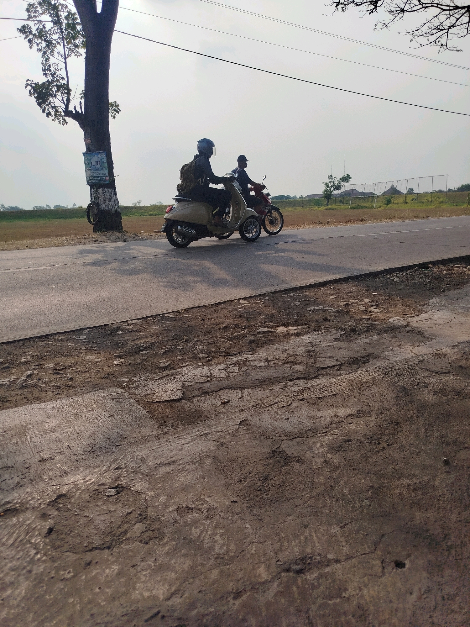 Two people on motorcycles are traveling on a road in Ngreco, Indonesia. The person in the foreground is on a light-colored scooter, wearing a dark helmet and a backpack. The person behind them is on a red motorcycle, also wearing a helmet. They are traveling on a paved road with a dirt shoulder. A large tree stands on the left side of the road with a poster attached to its trunk. In the background, there are fields, some buildings, and a fence. The sky is overcast.