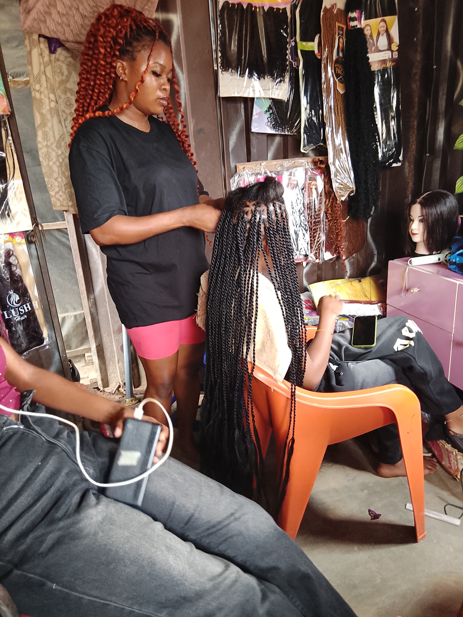 In a bustling salon setting in Ihiagwa, Nigeria, a young woman with striking, voluminous auburn braids styled into a high ponytail is intently braiding another person's hair. She wears a black t-shirt and vibrant pink shorts, her focus evident as she works with the dark braids. The client, seated in an orange plastic chair, has long, intricate dark braids extending down their back and wears a cream-colored top and dark track pants. They hold a black smartphone and a portable charger connected by a white cable, suggesting they are keeping themselves entertained or powered up during the braiding session.
The background of the salon is filled with various hair products, including neatly packaged bundles of synthetic hair in different colors and textures, suggesting a well-stocked establishment catering to diverse styling needs. A mannequin head with dark hair sits on a pink cabinet, further indicating the nature of the business. The lighting appears to be from artificial sources, consistent with an indoor setting, and there are no clear indicators of the time of day or weather outside. The overall atmosphere seems relaxed and productive, with the focus on the skillful artistry of hair braiding. Visible text includes "LUSH Hair" on a package of hair products to the left of the frame.