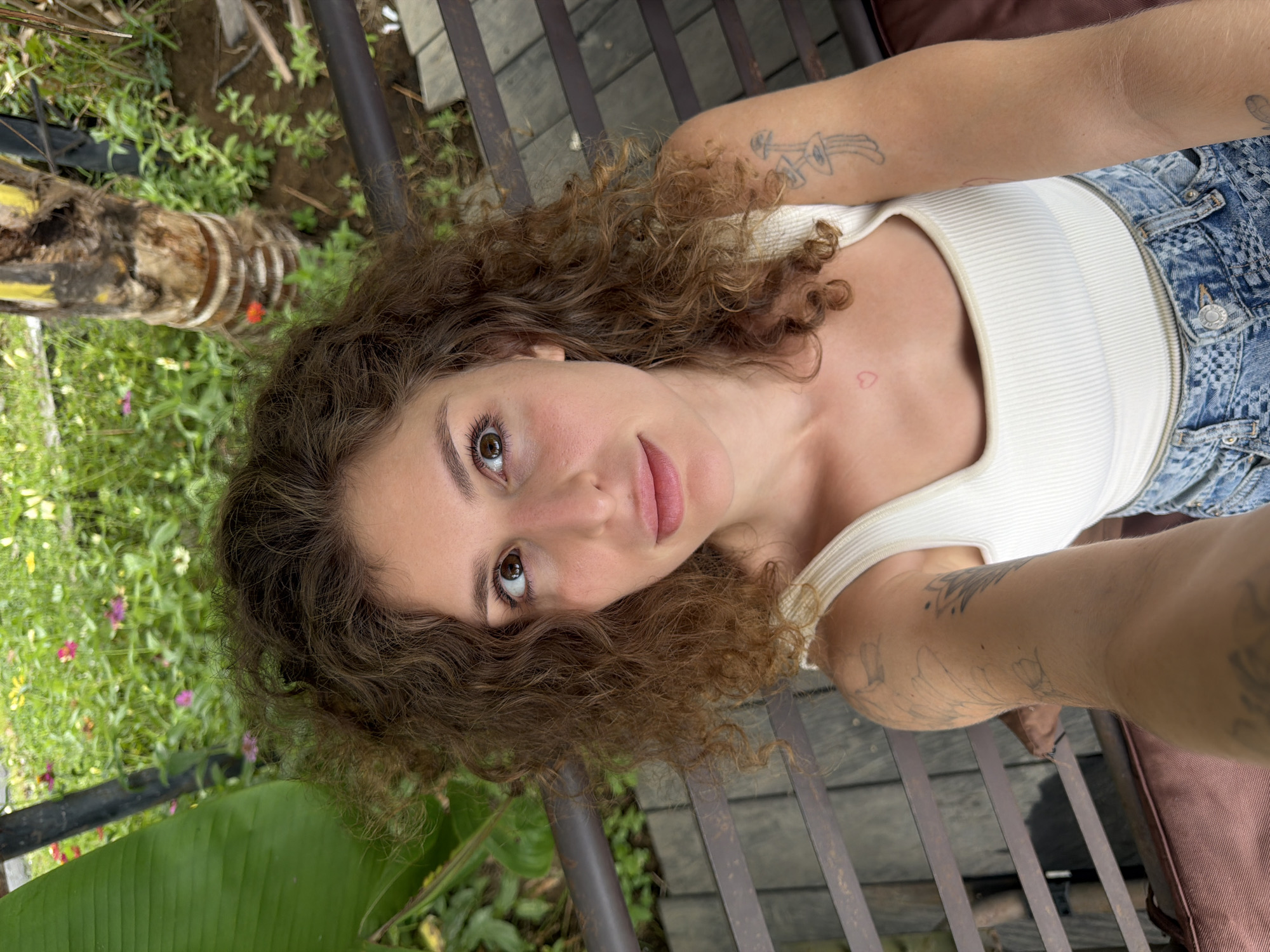 A young woman with curly brown hair is captured in a selfie taken from a high angle. She is seated on a dark brown slatted bench outdoors, with a blurred green background of plants and flowers. She is wearing a white ribbed tank top and blue denim jeans. Tattoos are visible on her right arm and a small heart tattoo is on her upper chest. She is looking upwards and to her right, with her mouth slightly open. The overall impression is a candid moment in a natural, outdoor setting.