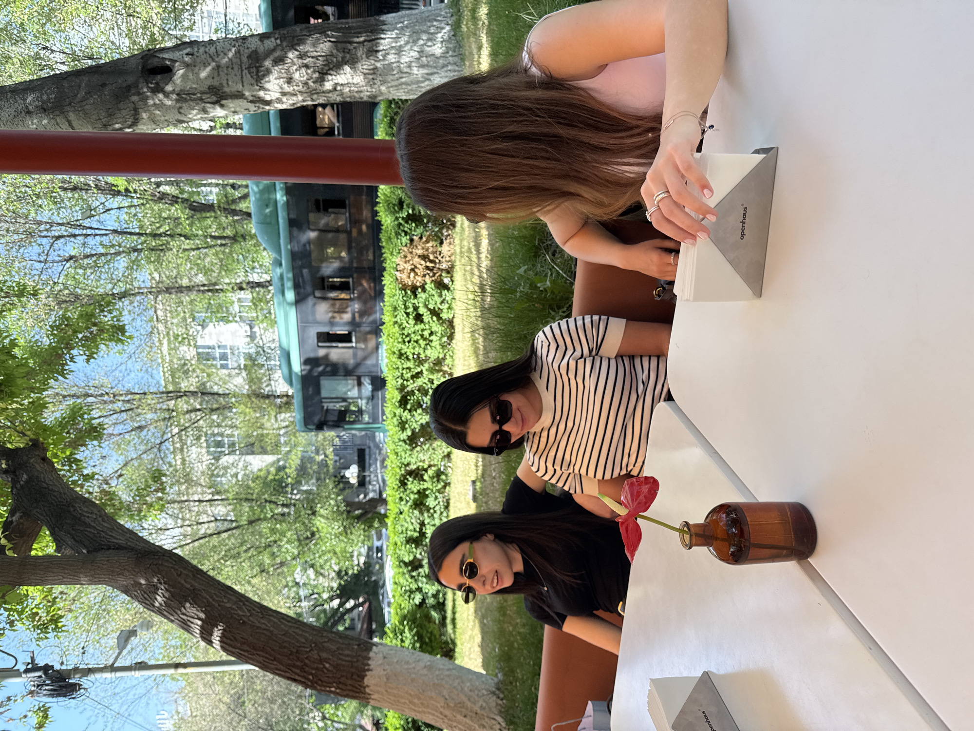 Three women are seated around a white table in an outdoor setting, likely a restaurant or cafe, potentially in Yerevan, Armenia. One woman, with light brown hair, is positioned at the top of the frame, her hands visible near a napkin holder labeled "openhaus." To her left, is another woman wearing a striped shirt and sunglasses. Further down is a third woman in a black shirt and sunglasses. A small brown vase containing a red flower sits on the table. In the background, lush greenery and trees are visible. A painted red metal structure and a vintage railway car add further detail to the scene.