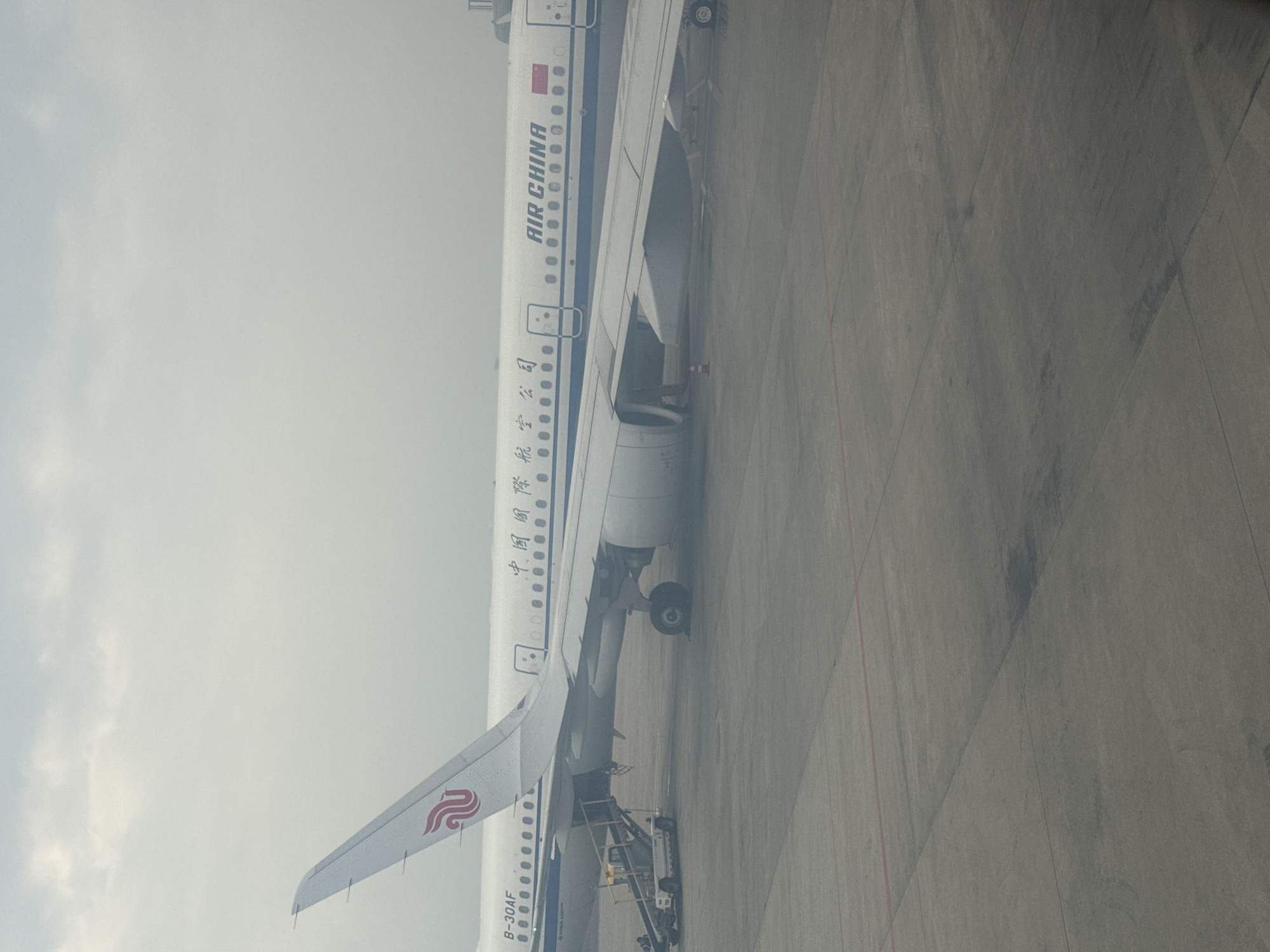 The image shows an Air China plane parked on the tarmac in Shuangliu District, China. The plane has its landing gear down, and a set of stairs is positioned next to it. The sky is overcast, and the ground is a grey concrete. The plane's tail is visible with the airline's logo.