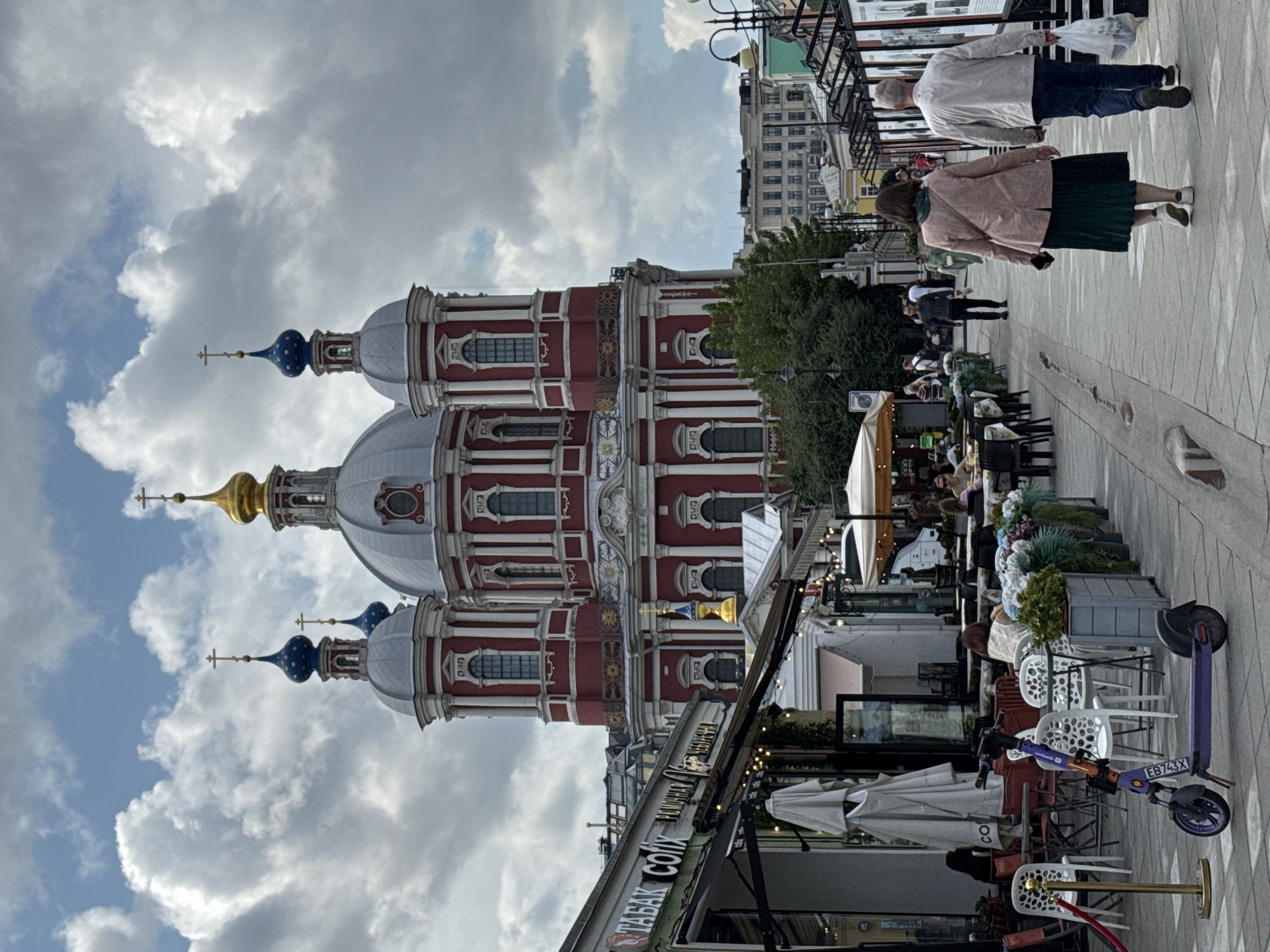 A vertical, eye-level, medium shot captures a lively street scene in Moscow, Russia, with a prominent red and white domed church as a backdrop. The church, adorned with blue and gold accents, features multiple domes and crosses. In the foreground, a cobblestone walkway is lined with outdoor seating areas of cafes and shops, indicated by tables, chairs, umbrellas, and signage with Russian text. Several people are visible, including a couple walking away from the camera, a woman in a pink jacket and green skirt, and a man in a light jacket and jeans carrying a white plastic bag. Other individuals are seated at tables, some conversing. A purple electric scooter is parked on the walkway. The sky is filled with dramatic, grey and white clouds, suggesting an overcast day. The environment appears to be a pedestrian-friendly area with a blend of historical architecture and modern amenities.