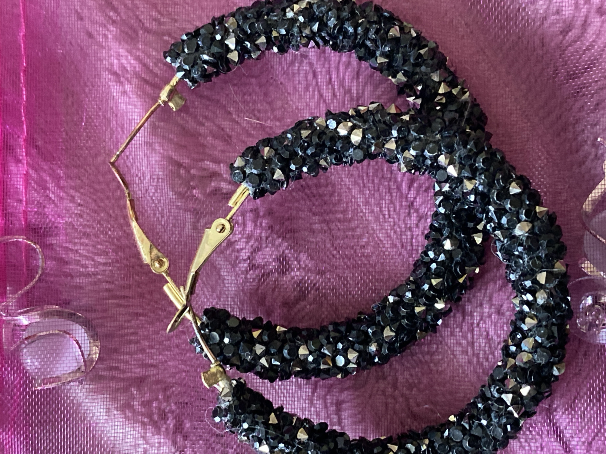 The image displays a pair of large, hoop earrings, primarily black in color, set against a vibrant fuchsia or deep pink fabric background. The earrings are thickly encrusted with numerous small, faceted beads or rhinestones, which give them a sparkling, textured appearance. Some of these embellishments appear to be black, while others reflect light with a metallic, silver-toned sheen. The earrings are held together by golden-colored hook-and-loop clasps, which are visible in the top and bottom sections of the image.
The background fabric has a noticeable weave or texture, appearing slightly crinkled and shiny, possibly satin or silk. In the bottom-left corner of the frame, a fragment of another object, silver-toned and possibly metallic or clear, is visible, suggesting a decorative element or another piece of jewelry.
The scene is a close-up, still-life shot of the jewelry, likely taken indoors. There are no people, explicit activities, visible text, or discernible time of day. The lighting emphasizes the glitter and texture of the earrings. No specific visual cues link the image to Novokuznetsk, Russia.