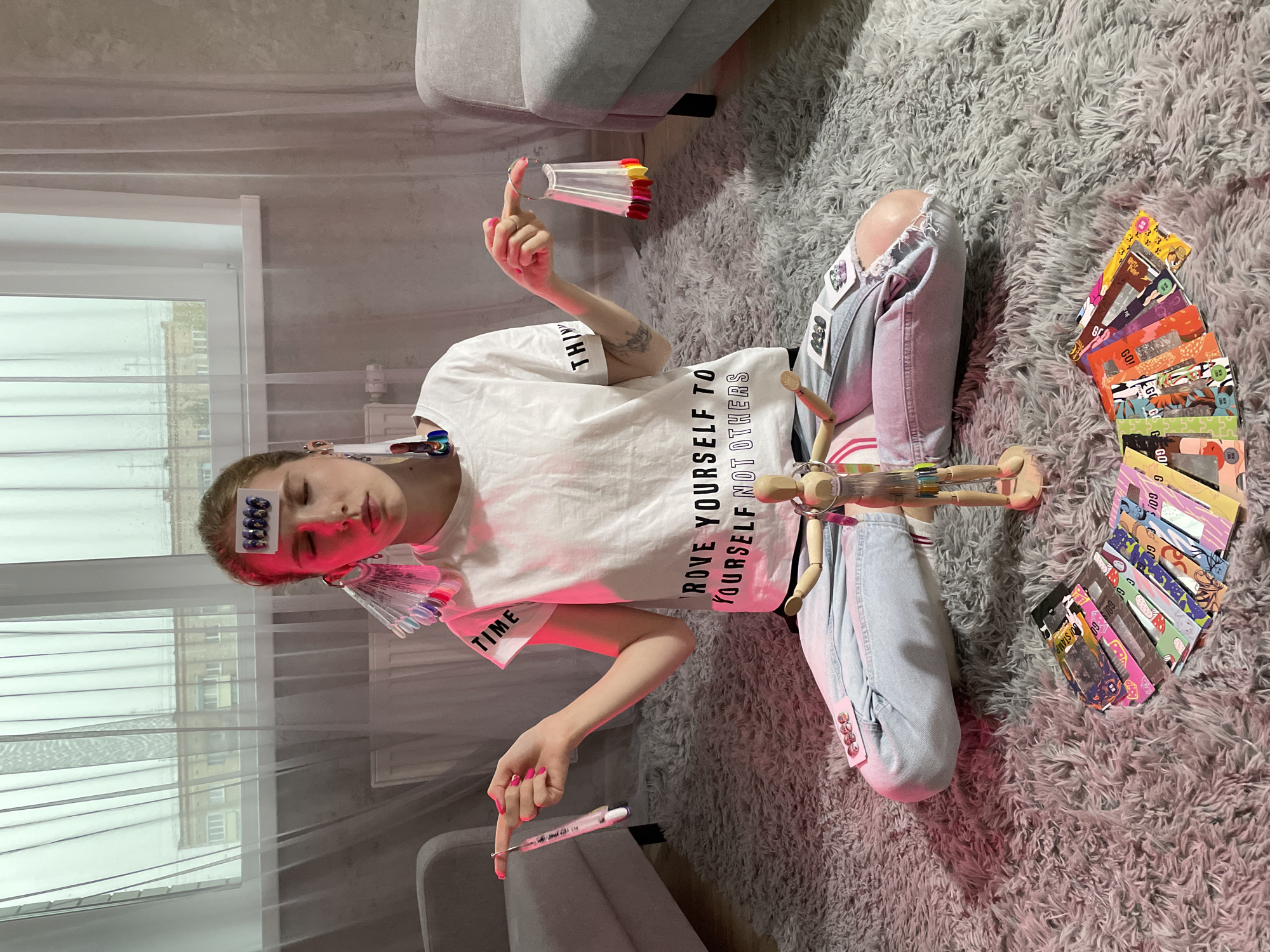 The image shows a woman sitting cross-legged on a grey carpet in an indoor setting. She wears light blue jeans and a white t-shirt with the text "IMPROVE YOURSELF TO YOURSELF NOT OTHERS" and "TIME". She has various items attached to her head and body, including nail polish samples, pill strips, and what appear to be banknotes. She holds a pen in one hand and a fan of nail polish samples in the other. A wooden mannequin lies in front of her. To the right of the woman is a fan of "GO! STAMP" cards. The location is identified from the text on the cards as Izhevsk, Russia. Through the window, an urban environment can be seen.