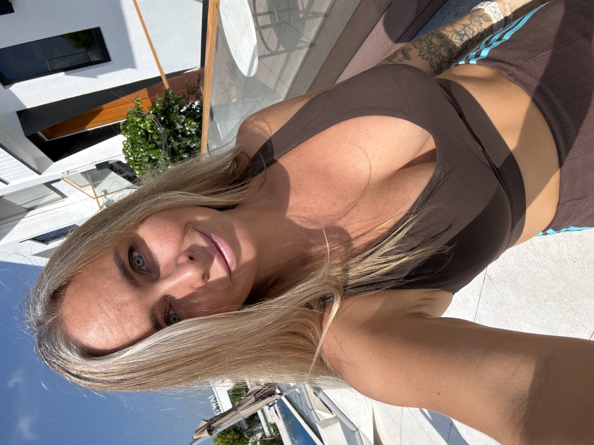 The image is a selfie taken by a blonde-haired woman outdoors. She is wearing a brown sports bra and brown athletic pants with light blue stripes. She has blue eyes and fair skin, with some gray streaks in her hair. She is smiling and looking at the camera. In the background, there is a modern building with white walls and dark-framed windows. There is also a green bush and a railing. To the left of the frame, a swimming pool and lounge chairs are visible, suggesting a resort or residential area. The overall lighting indicates a sunny day. The location of Pudong, China is not explicitly visible in the image.