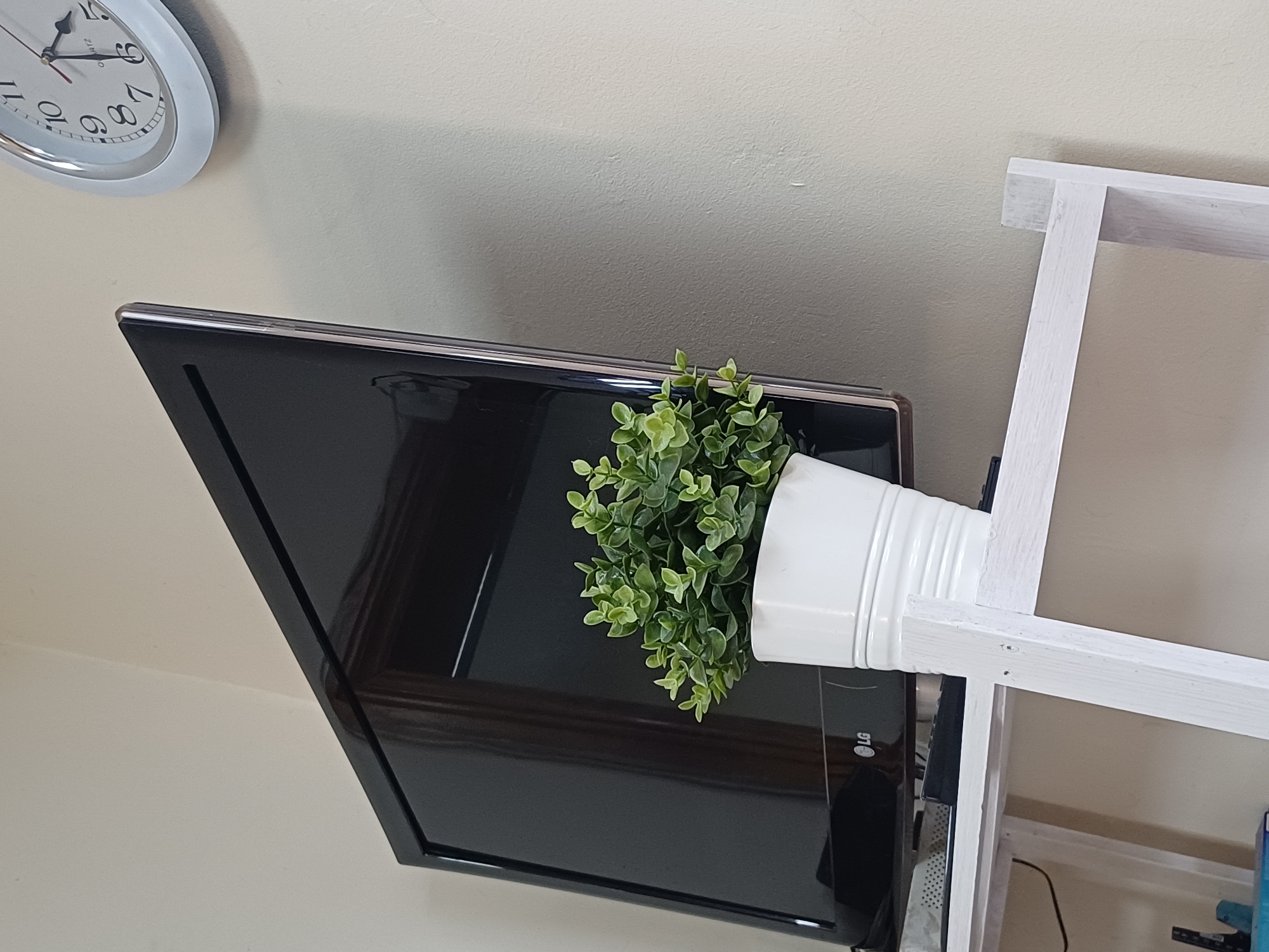 A television set is mounted on a wall, with a small potted plant placed in front of it. To the right of the television, a round wall clock with a white frame and black numerals is visible. The scene appears to be an indoor setting, possibly a living room or office, with a neutral-colored wall.