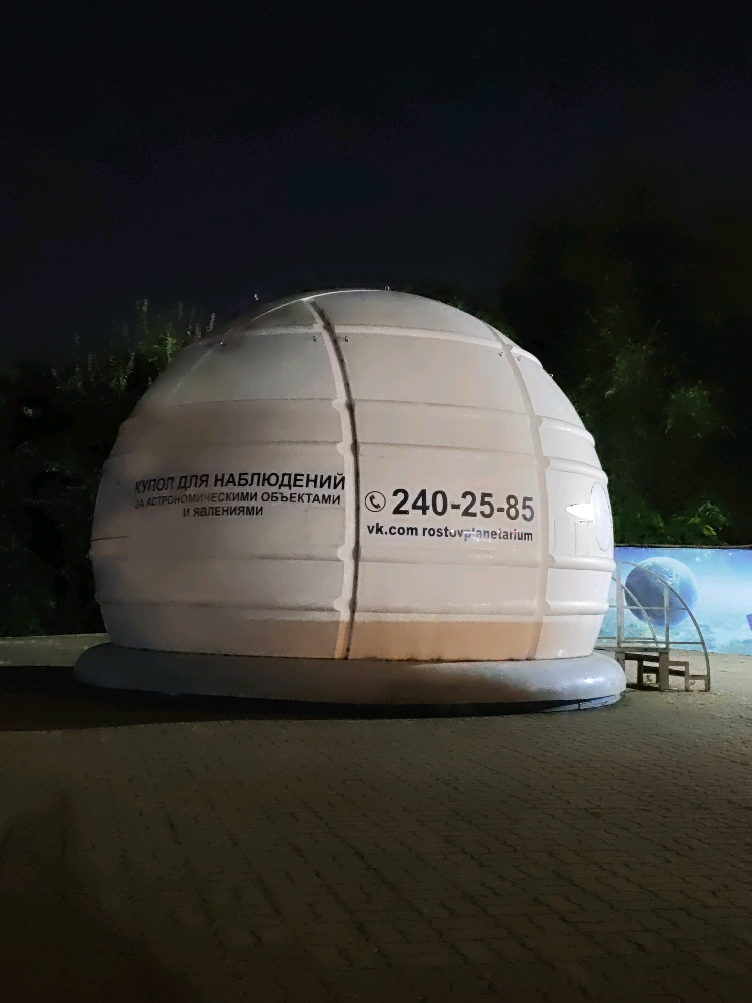 The image depicts a white, dome-shaped structure, likely a planetarium or observatory dome, situated outdoors at night. The dome has text printed on it in Russian. The text on the left side reads "КУПОЛ ДЛЯ НАБЛЮДЕНИЙ" (Dome for observations) and below that, "ЗА АСТРОНОМИЧЕСКИМИ ОБЪЕКТАМИ И ЯВЛЕНИЯМИ" (for astronomical objects and phenomena). On the right side of the dome, a phone number "240-25-85" is displayed along with a website address "vk.com/rostovplanetarium". This website address strongly suggests the location is the Rostov-on-Don Planetarium in Russia. The dome is supported by a circular, light-colored base. In the background, there are dark trees and a mural or display featuring a planet and a starfield, with a metal staircase leading up to the dome. The ground in front of the structure appears to be paved with textured blocks, and the overall lighting suggests an evening or nighttime setting.