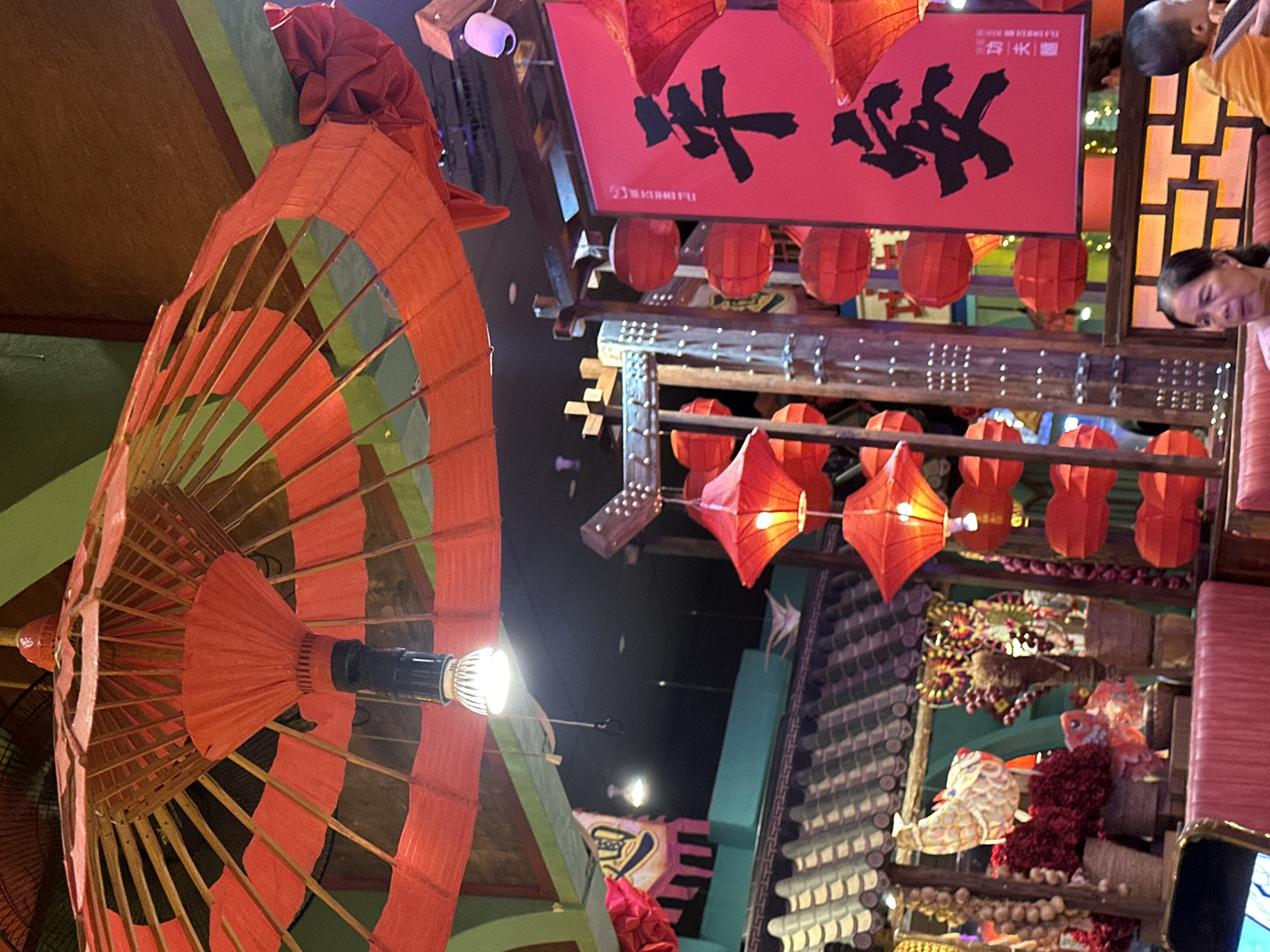 The media file depicts a vibrant indoor scene, likely a restaurant or market, decorated with traditional East Asian elements. A large, open red and green paper umbrella is prominently featured in the foreground, its bamboo frame and colorful paper visible. Above, strings of red lanterns hang, illuminating the space with a warm glow. A red banner with black calligraphic characters, possibly Chinese or Vietnamese, is displayed, with the text "福" visible on the right side. Several people are present, with their faces partially visible, suggesting a lively atmosphere. One person is looking down and to the left, while another's head is visible from the side. The setting appears to be dimly lit, with artificial lighting from the lanterns and some overhead fixtures. The overall impression is one of a festive and culturally rich environment. The presence of traditional decor and the context of Đà Nẵng, Vietnam, suggest a location that embraces and celebrates East Asian heritage.