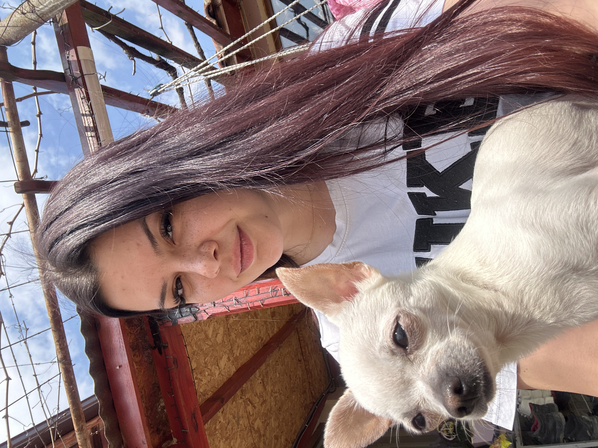 A woman with long, dark auburn hair is holding a small, white dog. The woman is wearing a white t-shirt with black print. They appear to be outside, under a structure with wooden beams and a metal roof. The sky is visible in the background. The photograph may have been taken in Chubra, Bulgaria.