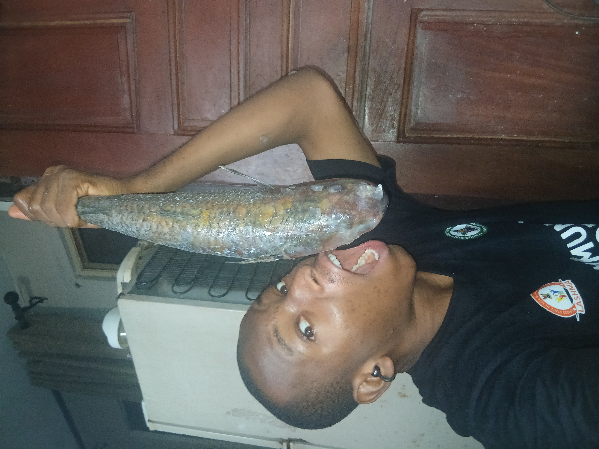 A young Black person is shown in a close-up shot, holding a large, silver fish with their right hand. The person's mouth is open, and they appear to be leaning in towards the fish, as if to bite it. The fish is long and slender, with visible scales and fins. The background is dimly lit, with a refrigerator visible to the left and a wooden door to the right. The person is wearing a black t-shirt with logos on it. The overall impression is one of playful or humorous interaction with the fish.