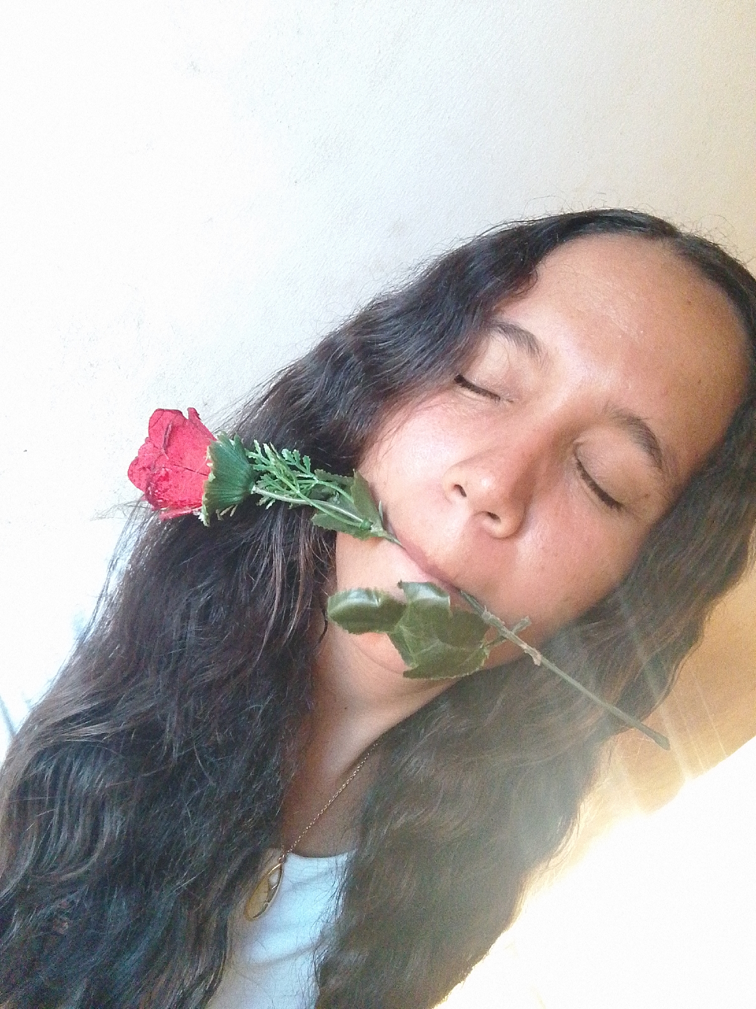 A close-up, tilted portrait features a young woman with long, dark, wavy hair. She is holding a red artificial rose by its stem in her mouth, with the red bloom positioned near her left ear. Her eyes are closed, conveying a sense of serenity or peacefulness. The woman has a light brown complexion and is wearing a white top with a delicate gold necklace and a light-colored pendant. The background is a plain, light-colored wall. Bright, possibly natural, light illuminates the scene, causing some overexposure in the bottom right corner of the image. The lighting suggests it is daytime. No text is visible in the image. The context for this image is Juan Griego, Venezuela.