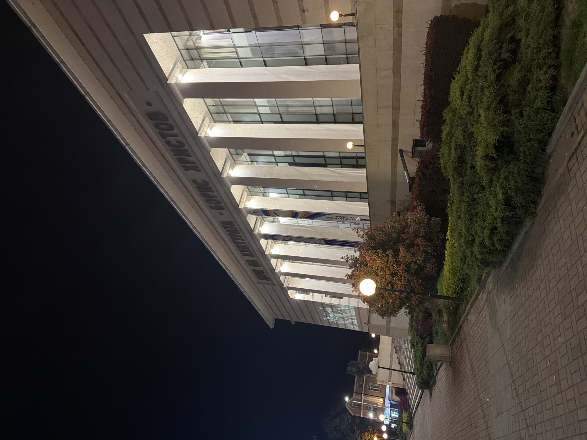 The image captures a nighttime view of a large building, likely an opera house, in Plovdiv, Bulgaria. The building's facade is illuminated, revealing multiple vertical columns and large windows that reflect some interior lighting. Cyrillic text is visible on the building's exterior, reading "ИМ. КИТНА БОРИС ХРИСТОВ" (IM. KITNA BORIS HRISTOV) on a prominent band, and part of a banner or sign through the windows reads "ОПЕРА ПЛОВДИВ" (OPERA PLOVDIV), confirming its identity as an opera venue.
In front of the building, a landscaped area features dense green bushes and some reddish-brown foliage, separating the structure from a paved pathway made of interlocking bricks. Several spherical street lamps with a warm, yellowish glow are distributed along the path, providing additional illumination to the scene. The sky is dark, indicating it is night, and there are no visible clouds. No people are present in the frame. The overall impression is one of a well-maintained public space in an urban setting at night.