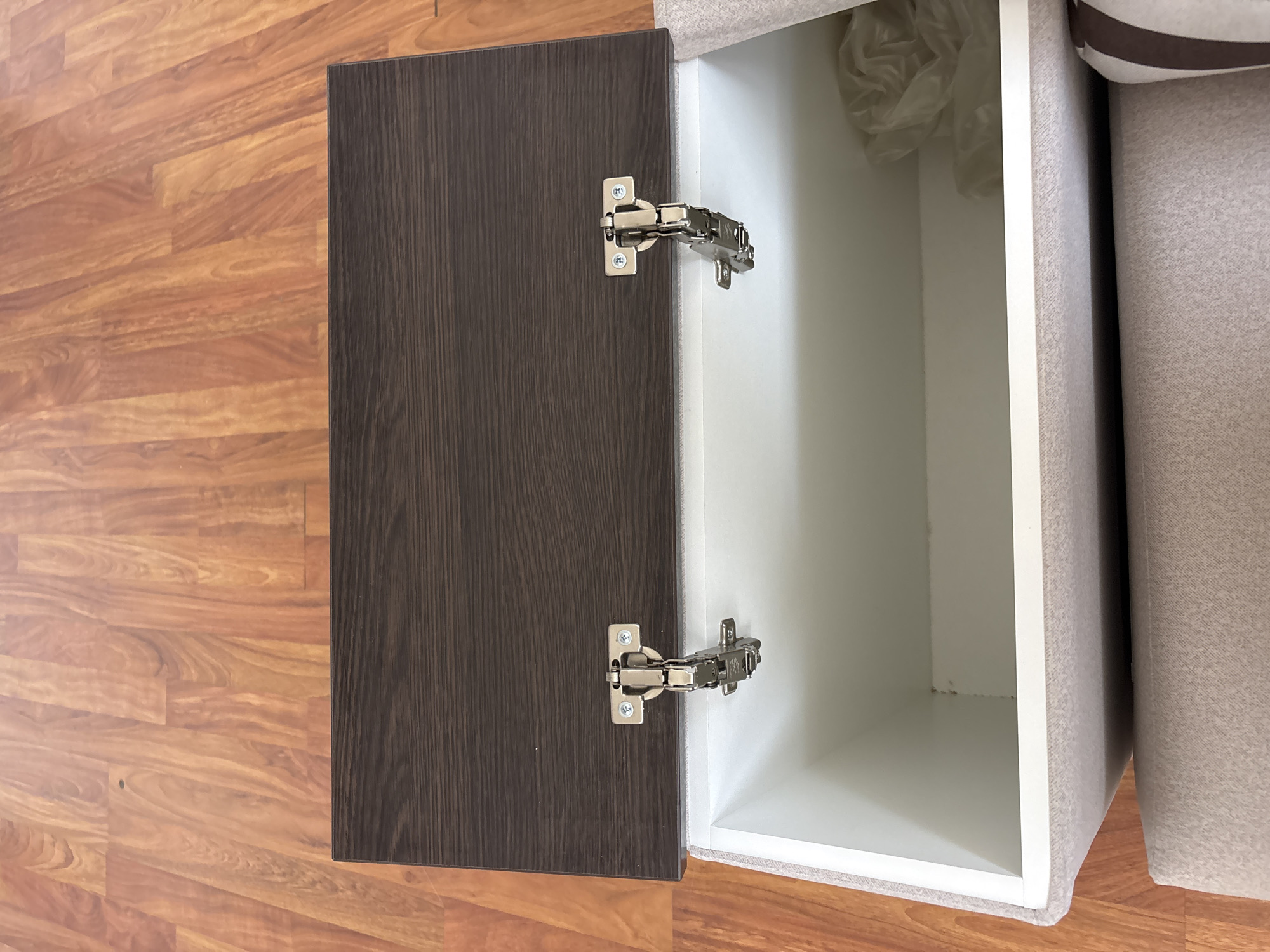 An open storage ottoman with a dark brown wooden lid rests on a light brown upholstered base. The ottoman is positioned on a light brown wooden floor. The lid, held open by two silver hinges, reveals the white interior of the ottoman, where a crumpled white plastic bag is visible. The scene is indoors, with no discernible outdoor environment or specific location within Zaporizhzhia, Ukraine.