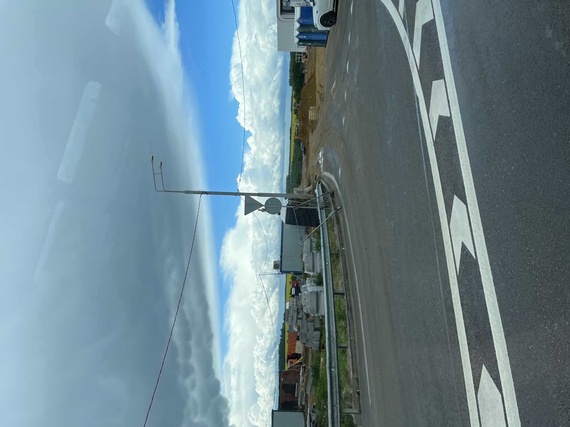 The image depicts a cloudy sky above Umryshenka, Russia, next to a construction area with parked portable restrooms and an unfinished asphalt road. There is a pile of sand, and a truck parked along the road. Construction materials are next to the road. A reflection of a window is visible.