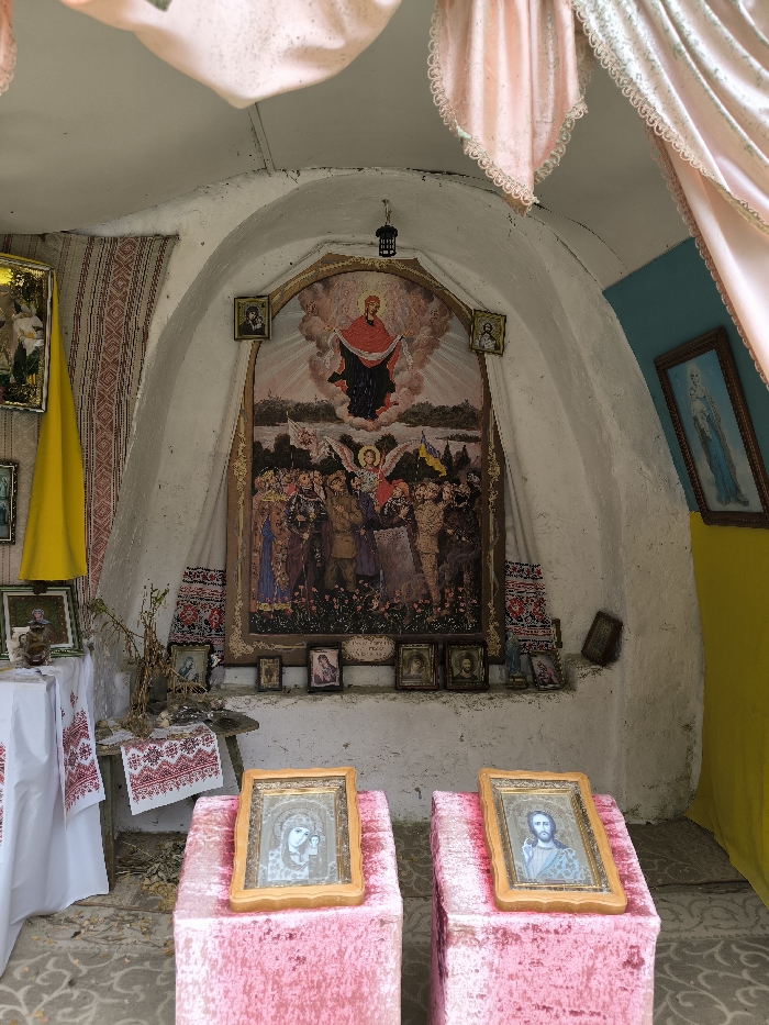 This image depicts the interior of a small, arched, white-washed structure, likely a chapel or shrine, located in Horaivka, Ukraine.
The central feature on the rear wall is a large, vertically oriented, gold-framed religious painting. The upper portion of this painting displays a figure in red and dark robes, possibly the Virgin Mary, suspended within clouds. The lower portion depicts a gathering of figures, including an angel with wings, individuals in military uniforms (some appearing contemporary), and others in civilian attire, standing in a field. Two Ukrainian flags are visible within this crowd. Flanking this primary painting, at a higher elevation, are two smaller, framed religious icons. Below the main painting, a narrow ledge holds a horizontal row of multiple small, framed icons. A long, white textile with red embroidered patterns hangs vertically behind the central painting. A dark, lantern-like fixture is suspended from the arched ceiling directly in front of the main painting.
To the left of the central arrangement, a wooden table or bench is partially covered by a white textile with red embroidered designs. On this surface, dried plant material and other small objects are present. Above and to the left of the table, another framed image is partially visible behind a draped yellow fabric.
The right wall is divided horizontally, with a light blue upper section and a yellow lower section. A framed image of the Virgin Mary, depicted in blue tones, hangs on the upper blue section. A small, dark framed object leans against the wall below.
In the foreground, two rectangular pedestals, covered in pink, textured fabric, stand centrally. Each supports a framed religious icon tilted towards the viewer. The icon on the left depicts the Virgin Mary holding the infant Jesus. The icon on the right depicts Jesus Christ. Pink, ruffled textiles also drape from the upper curved ceiling sections on both the left and right. The floor in the foreground displays a patterned surface.