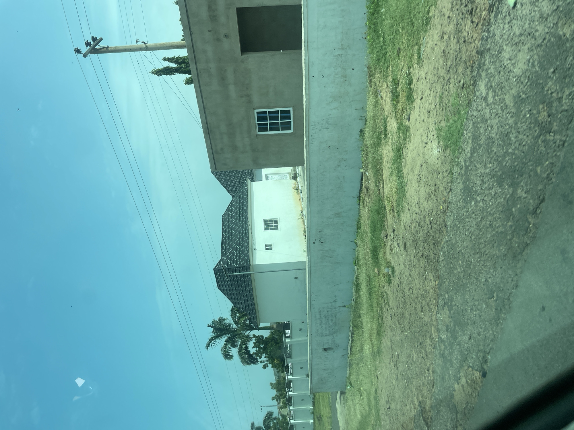 The image captures a residential scene in Chum, Nigeria, on a clear day. In the foreground, a rough, grey asphalt road with scattered patches of green grass and dirt occupies the lower portion of the frame. A light grey concrete wall runs horizontally across the mid-ground, appearing somewhat weathered and showing faint graffiti. Behind this wall, a modern-style white house with a dark, angular roof and a white-framed window is visible. To the right of the white house, another building is under construction; it is characterized by unfinished, tan-colored concrete walls, a dark, open doorway, and a white-framed window. A tall palm tree stands behind the white house, and other greenery, including trees and shrubs, are present. Overhead, a utility pole with wires stretches across the bright blue sky. The overall impression is of a developing residential area.