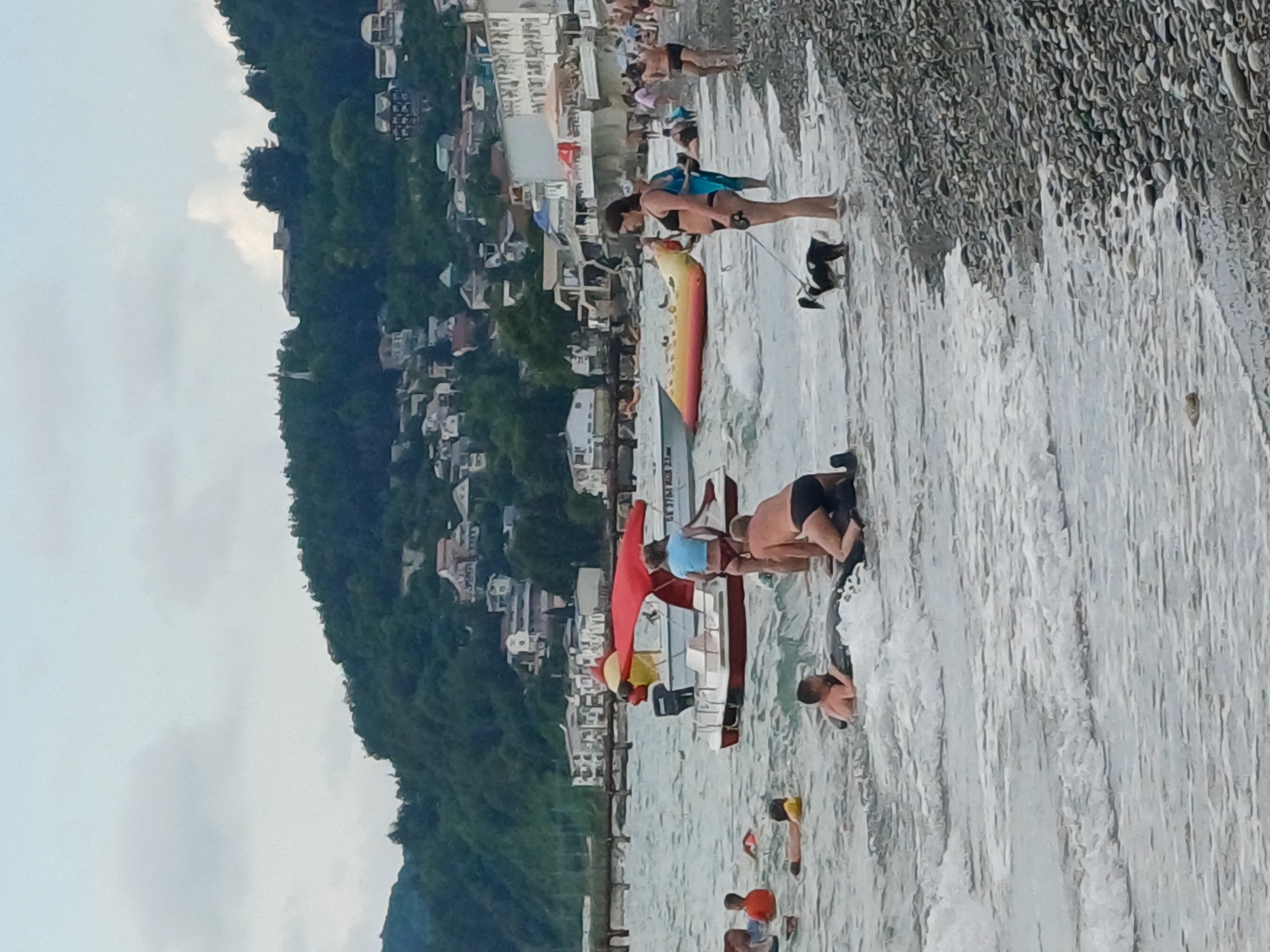 The image depicts a crowded beach scene in Sochi, Russia. The foreground is dominated by the ocean waves crashing onto a pebbly shore. Several people are in the water, some wading and others appearing to be swimming or playing. In the middle ground, there are more people on the beach, some sunbathing and others engaged in various activities. A colorful inflatable watercraft, possibly a surfboard or a small raft, lies on the sand.
Behind the beach, a steep, densely forested hillside rises, dotted with numerous buildings and houses, suggesting a densely populated coastal area. The architecture of the buildings hints at a Mediterranean or similar style. The sky is overcast, indicating a cloudy day, and the time of day appears to be afternoon or early evening, based on the light.
One person in the middle ground, a woman wearing a black bikini, is visible walking a small black dog on a leash. Another individual in the mid-ground is wearing a blue shirt and appears to be interacting with someone on the inflatable. Further back on the beach, more individuals are visible, many in swimwear. The overall atmosphere is one of a busy holiday resort, with many people enjoying the beach and the sea. There is no visible text in the image.