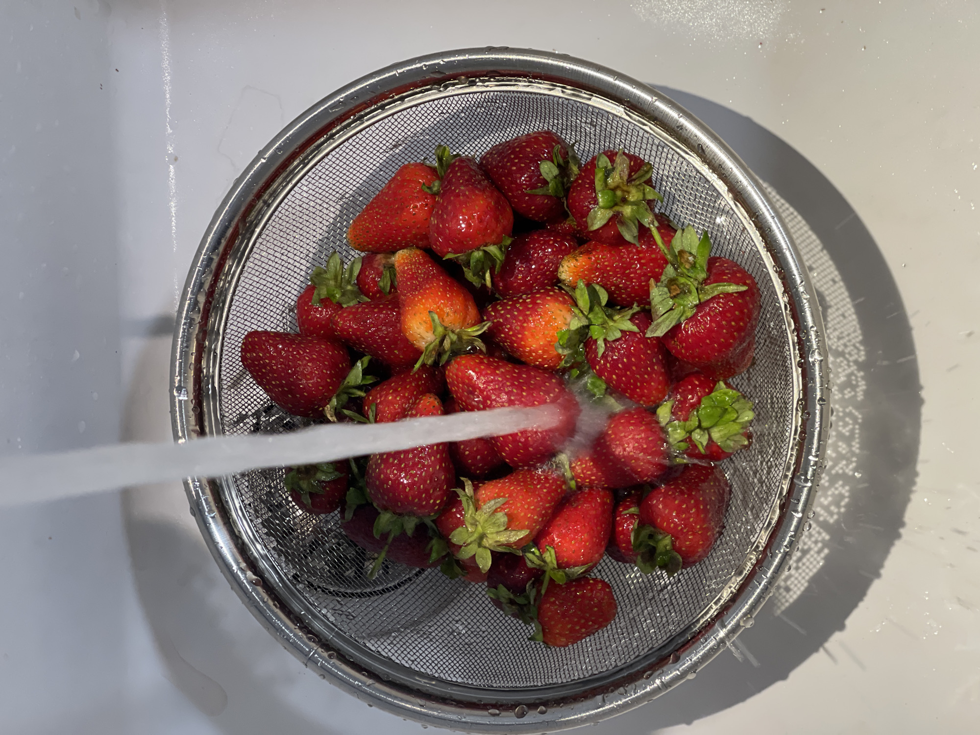 A top-down view shows a metal strainer filled with fresh strawberries being rinsed under a stream of water in a white sink. The strawberries vary in ripeness, from deep red to lighter orange-red, and most have green stems attached. Water droplets are visible on the strainer and surrounding sink surface. The image was not necessarily taken in the city of Izhevsk, Russia.