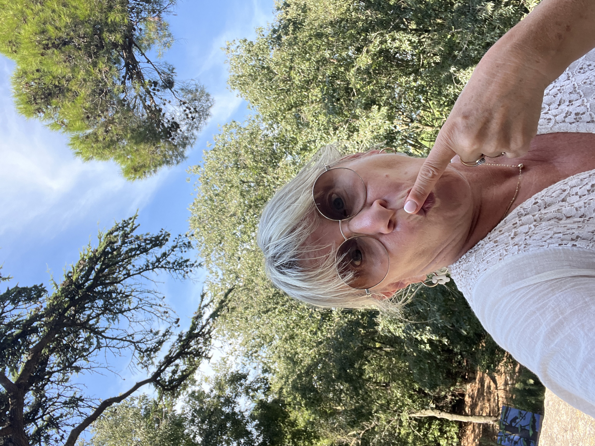 In Villeneuve-lès-Maguelone, France, a woman with blonde hair and sunglasses poses for a selfie outdoors. She wears a white, lace-trimmed top and a gold necklace. Her right index finger is raised to her lips, and she appears to be making a kissing gesture. The background is filled with lush green trees and a bright blue sky with scattered clouds.