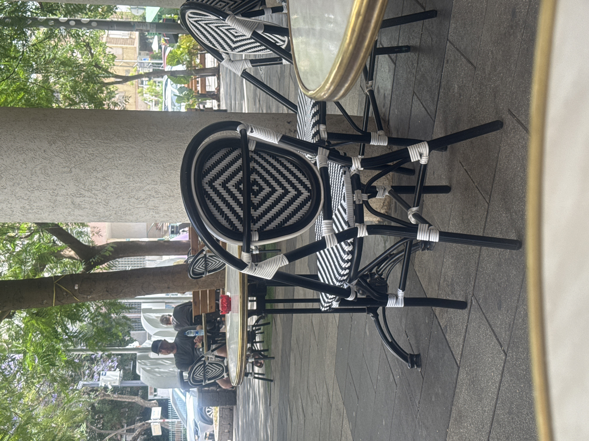 The image shows a sidewalk cafe in Givatayim, Israel. The cafe furniture is black metal with woven black and white seats and a round table with a gold rim. A man wearing a baseball cap is sitting at a table. A woman is visible behind him. Trees with green foliage frame the scene. A street with cars and buildings is visible in the background.
