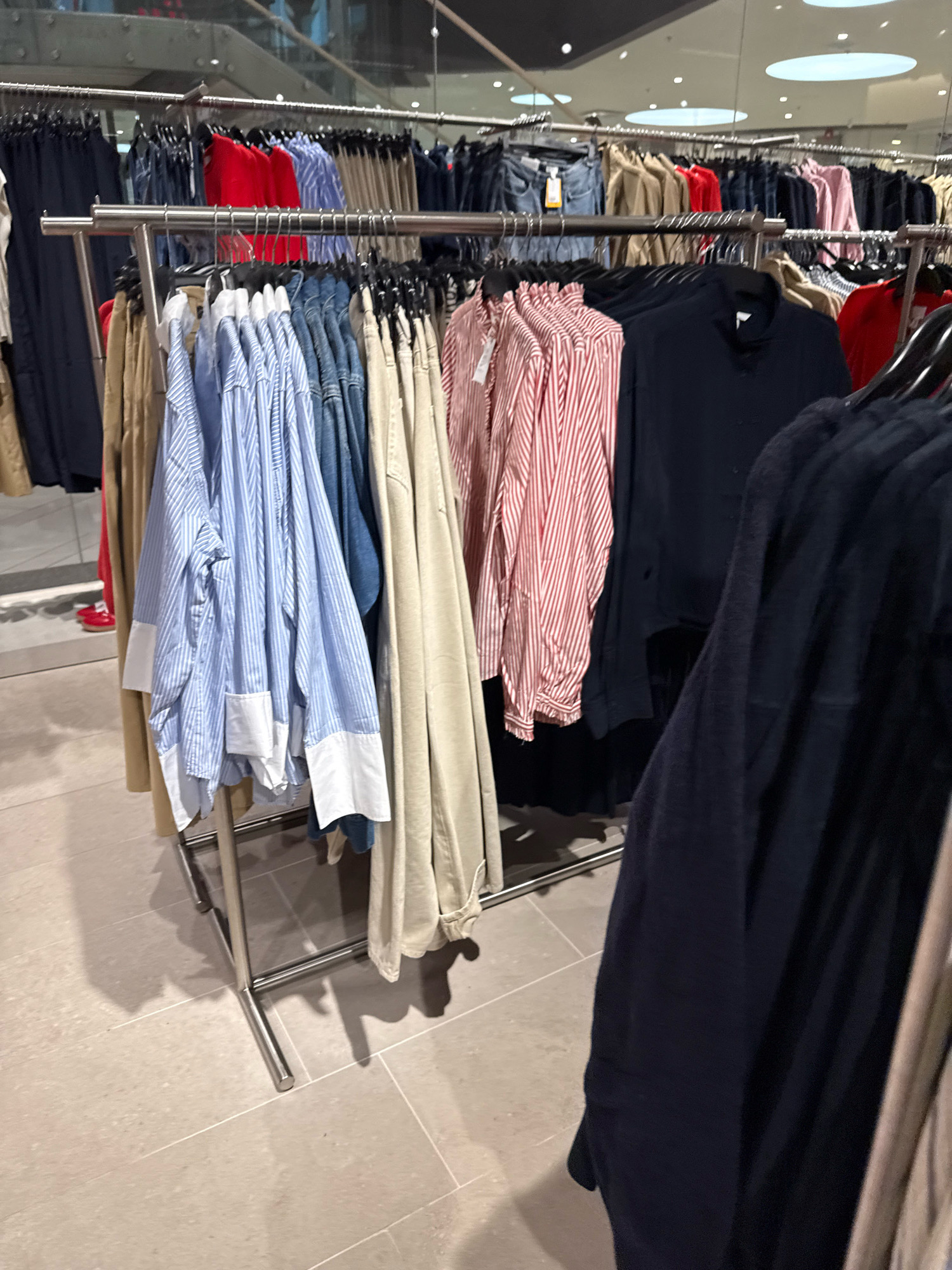 The image depicts a clothing store in Tallinn, Estonia. The scene focuses on clothing racks filled with various garments, including striped shirts, denim, and solid-colored tops. The clothing is neatly arranged and displayed on hangers. In the background, escalators and elevated walkways suggest a multi-level retail environment. The lighting is bright and even, typical of a commercial space. The floor is tiled with large, light-colored rectangular tiles. There are no discernible people in the immediate foreground, but a pair of red shoes is visible in the reflection of a mirrored surface on the left side of the image.