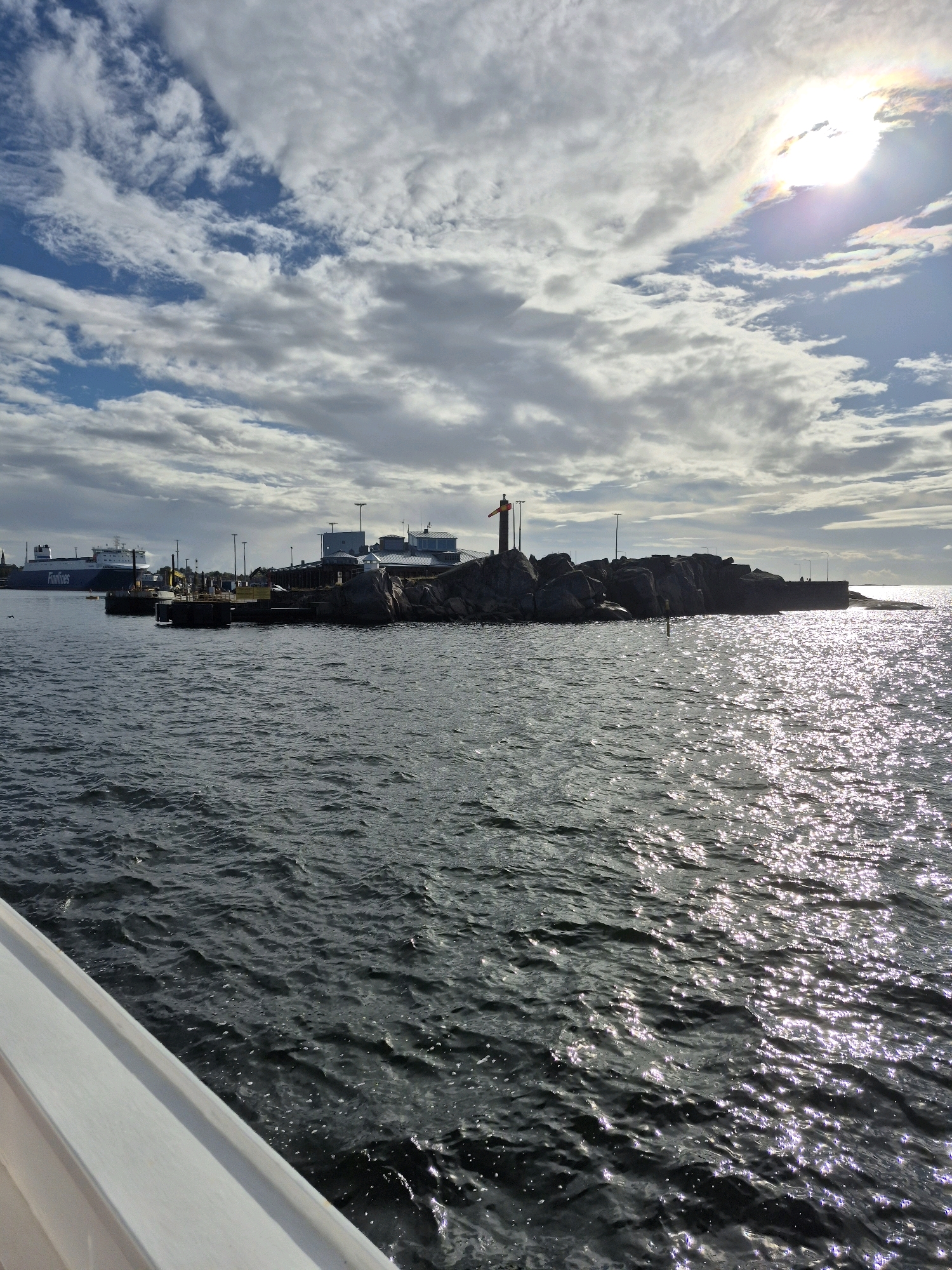 The image shows a view of the harbor in Hanko, Finland. In the foreground, a white railing of a boat or ferry is visible on the left. The dark, choppy water of the harbor fills the lower half of the image, with sunlight glinting off its surface. In the mid-ground, a rocky island with a small statue of a person is situated in the water. Behind the island, several buildings with industrial features and some lights are visible on the shoreline, suggesting a port or industrial area. Further in the background, a large ferry with "Finnlines" written on its side is docked. The sky is partly cloudy, with patches of blue visible and the sun partially obscured by clouds, creating a bright, slightly hazy light. The overall scene depicts a harbor area with maritime activity under a dynamic sky.