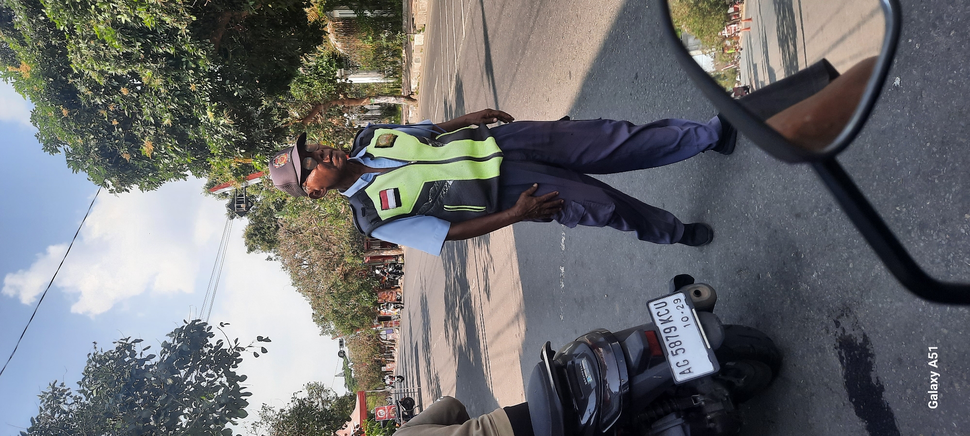 A full-shot, medium-distance, eye-level, outdoor, natural-light, daytime photograph shows an elderly Indonesian man in what appears to be a city street in Beru, Indonesia. The man is wearing a dark baseball cap with an emblem on the front, clear-rimmed glasses, a light blue collared shirt, a high-visibility vest with reflective green and gray accents, and dark gray cargo pants. The man's left hand is resting on his left hip, and his right hand is partially visible in front of him. The man is standing on an asphalt road, and a dark-colored scooter with a license plate reading "AG 5879 KCU 10-29" is parked to his left, partially in the foreground. The background shows a street with trees lining both sides, several shops or stalls, and other vehicles, including scooters, on the road. The sky is clear with some clouds. The scene suggests a warm, sunny day.