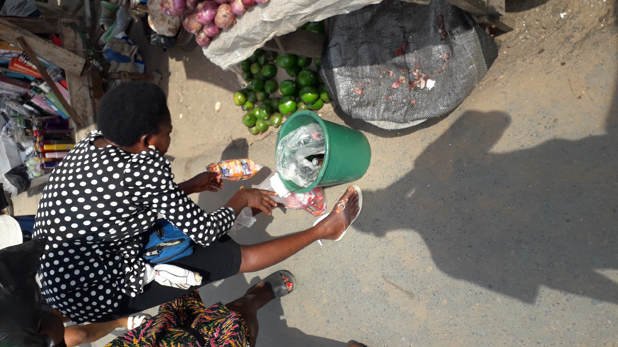 In a high-angle shot taken in Onitsha, Nigeria, a woman in a black and white polka dot dress sits on a dark-colored object, possibly a stool, alongside a paved street. She is seen handling packaged goods, possibly food items, with a green bucket filled with plastic bags and other goods next to her. Beside the bucket are scattered green, round fruits. To the woman's left is a pile of miscellaneous items, including books and other goods on a makeshift wooden stand. A sack of red onions is visible in the upper left corner of the image. A black mesh bag with what appears to be organic waste is next to the fruits. The woman is wearing a pair of white sandals, while another person’s feet are visible at the bottom of the image, wearing colorful printed clothing and sandals. The pavement, likely a road or sidewalk, is a medium-toned gray and has a large shadow cast upon it.