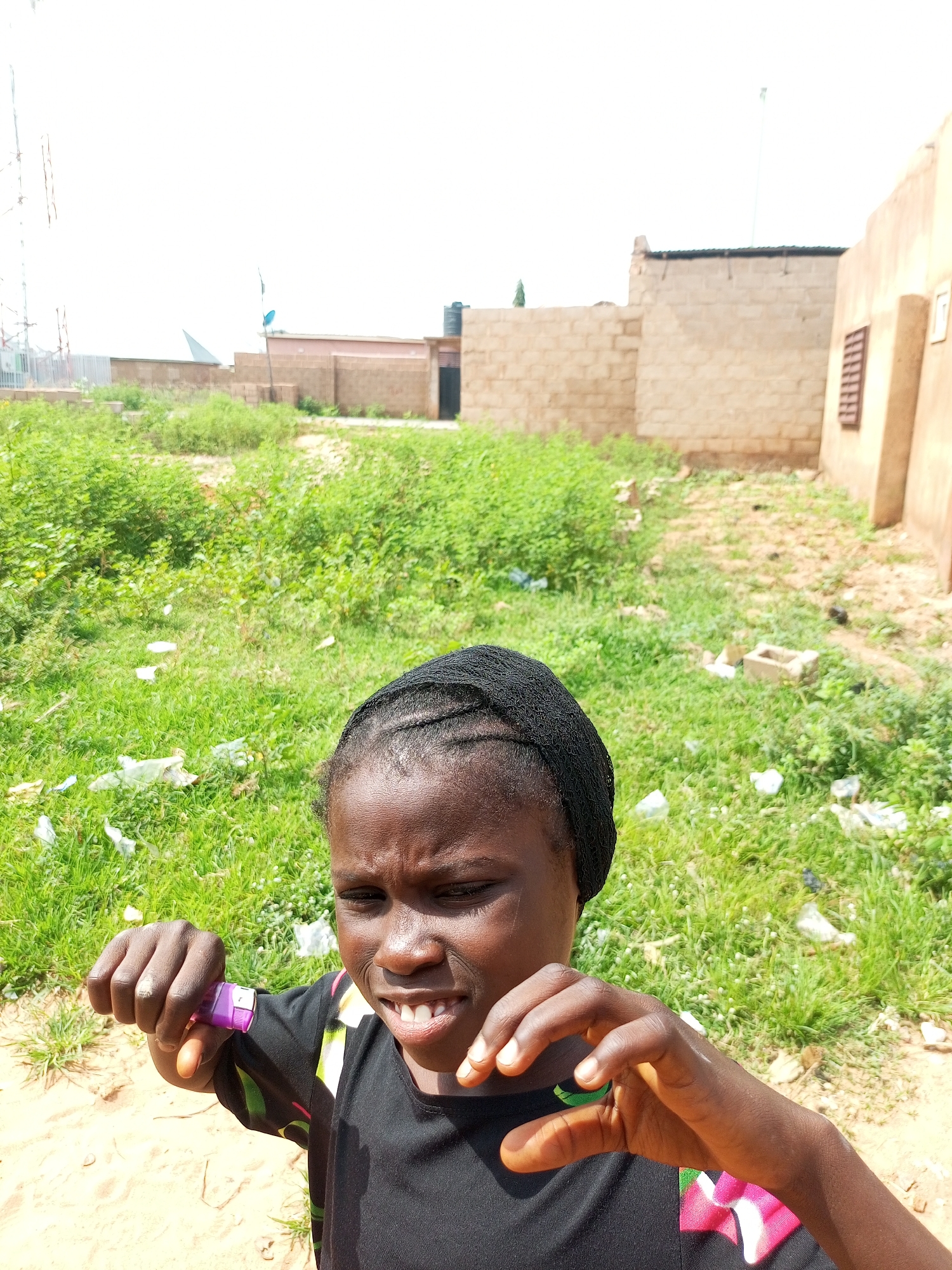 A young girl wearing a black headscarf and a black t-shirt with colorful details stands in a grassy, sandy area. Her hands are raised, and she is holding a small purple object in her left hand. She is looking towards the camera with a slightly scrunched facial expression, her teeth visible. The background consists of green grass with scattered white plastic trash. In the distance, there are low-rise buildings made of light-colored brick under a bright, overcast sky. A tall metal structure, possibly an antenna or transmission tower, is visible on the left side of the frame. The scene appears to be set outdoors during the daytime. There is no visible text in the image.