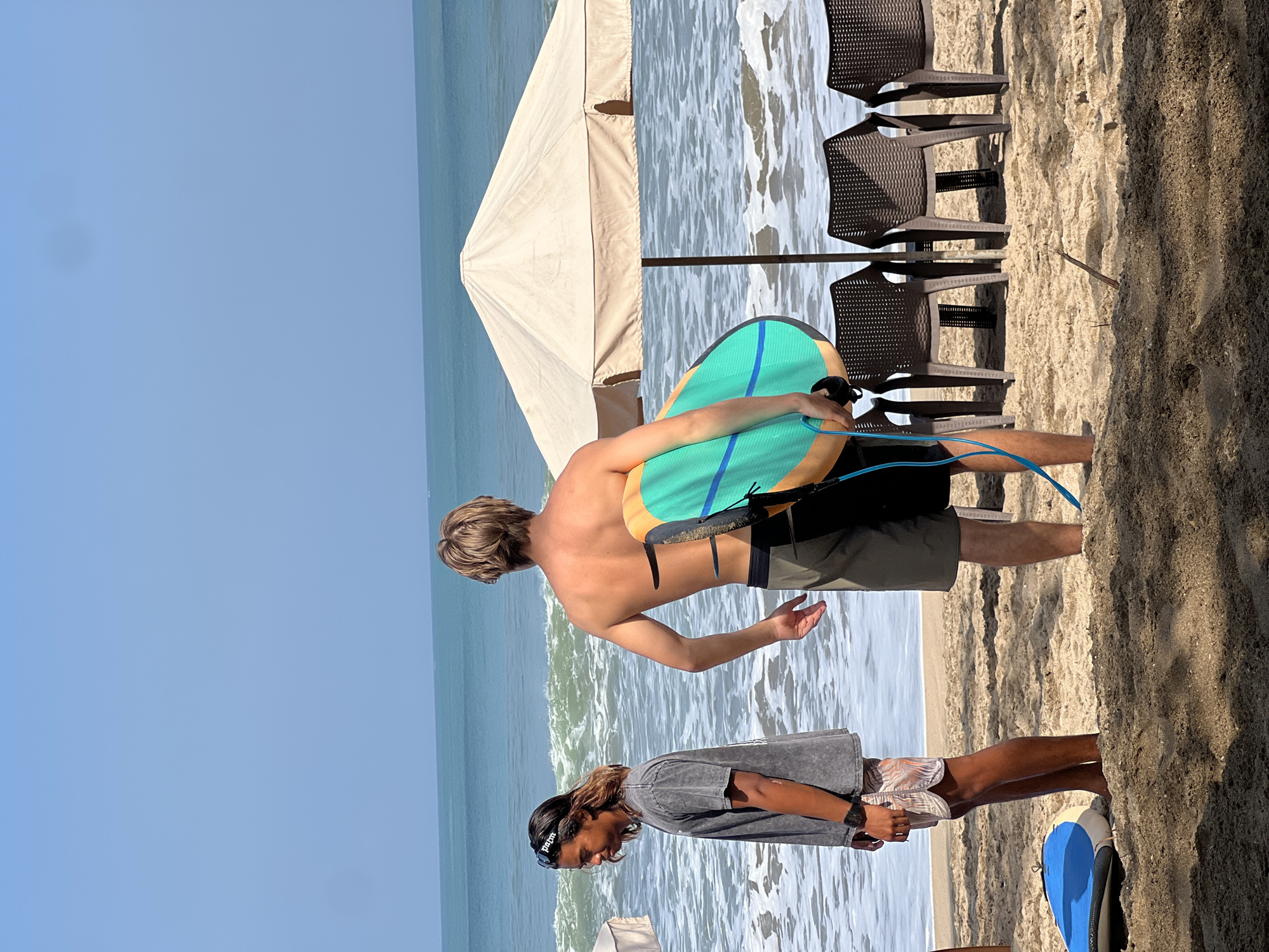 Two young men are on a sandy beach in Kuta, Indonesia, with the ocean in the background. The man on the left, wearing a grey t-shirt and patterned shorts, stands facing left, looking down. He has dark hair and is wearing sunglasses on his forehead. The man on the right, with blond hair, stands facing away from the camera, holding a turquoise and orange surfboard tucked under his right arm. He is wearing dark shorts. Behind the men, a large, light-colored beach umbrella shades several empty plastic chairs. The ocean waves are visible, and the sky is clear blue.
