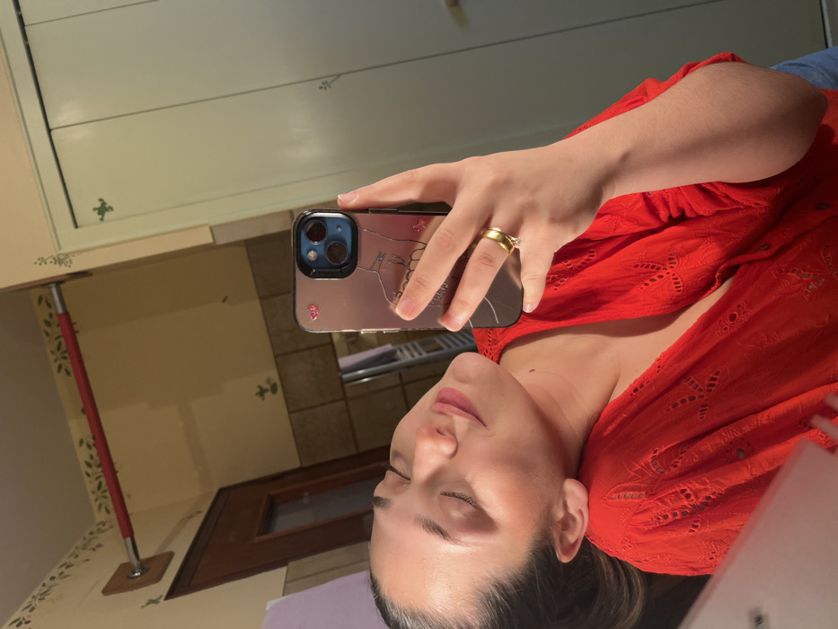 An image, identified as taken in Ancenis, France, depicts a mirror selfie of a female individual. The individual, positioned off-center to the left, has dark hair pulled back and wears a red, short-sleeved top with eyelet embroidery. Her eyes are closed, and her head is tilted slightly back and to the right, exhibiting a subtle smile or serene expression. She holds a smartphone in her right hand, reflected in the mirror. The phone case is metallic-toned, shows a prominent crack, and features a minimalist line art drawing of an upper body, along with small pink butterfly stickers near the camera module. A gold-colored band with a single clear stone is visible on her right ring finger. The background, also a reflection, reveals an interior space, likely a bathroom. The walls are light beige with a green stencil pattern depicting small floral or leaf motifs. A red horizontal bar is mounted on the upper left wall, secured by a wooden backplate. To the right, a light-colored, possibly green or white, two-panel door or cabinet with similar stencil details is visible. A metallic, ladder-style fixture, consistent with a towel warmer or radiator, is partially visible in the lower central reflected area.
