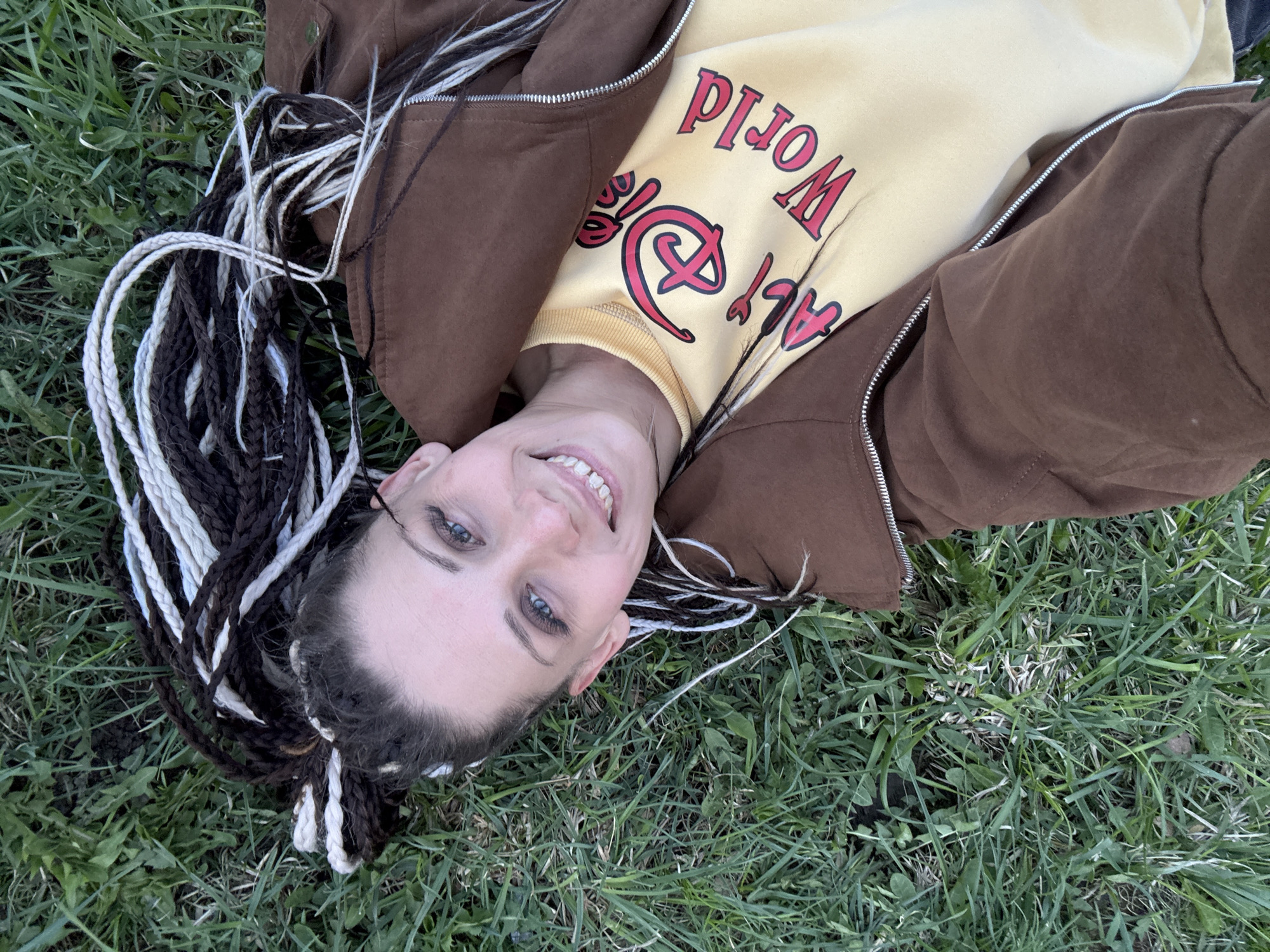 A fair-skinned person is lying on their back in a grassy area. They are wearing a yellow sweatshirt with red lettering, partially obscured by an unzipped brown jacket. The person has dark and light-colored braids. The location is a grassy area, possibly a park, but not identifiable as Rusanovo, Russia. The person is smiling and looking toward the camera.