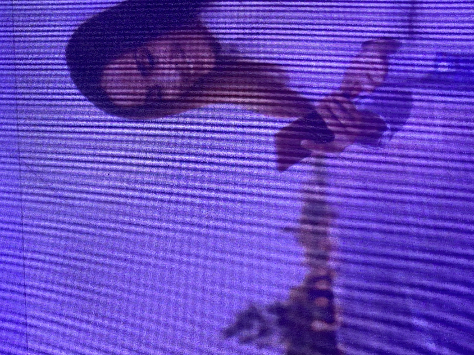 The image shows a woman standing outdoors, looking at her smartphone. She is wearing a white collared shirt. The background consists of blurred trees and buildings, suggesting an urban environment, potentially in Ikorodu, Nigeria, given the context. The overall color tone of the image is predominantly purple, possibly due to lighting conditions or camera settings. The woman appears engaged with her phone, with a slight smile on her face.