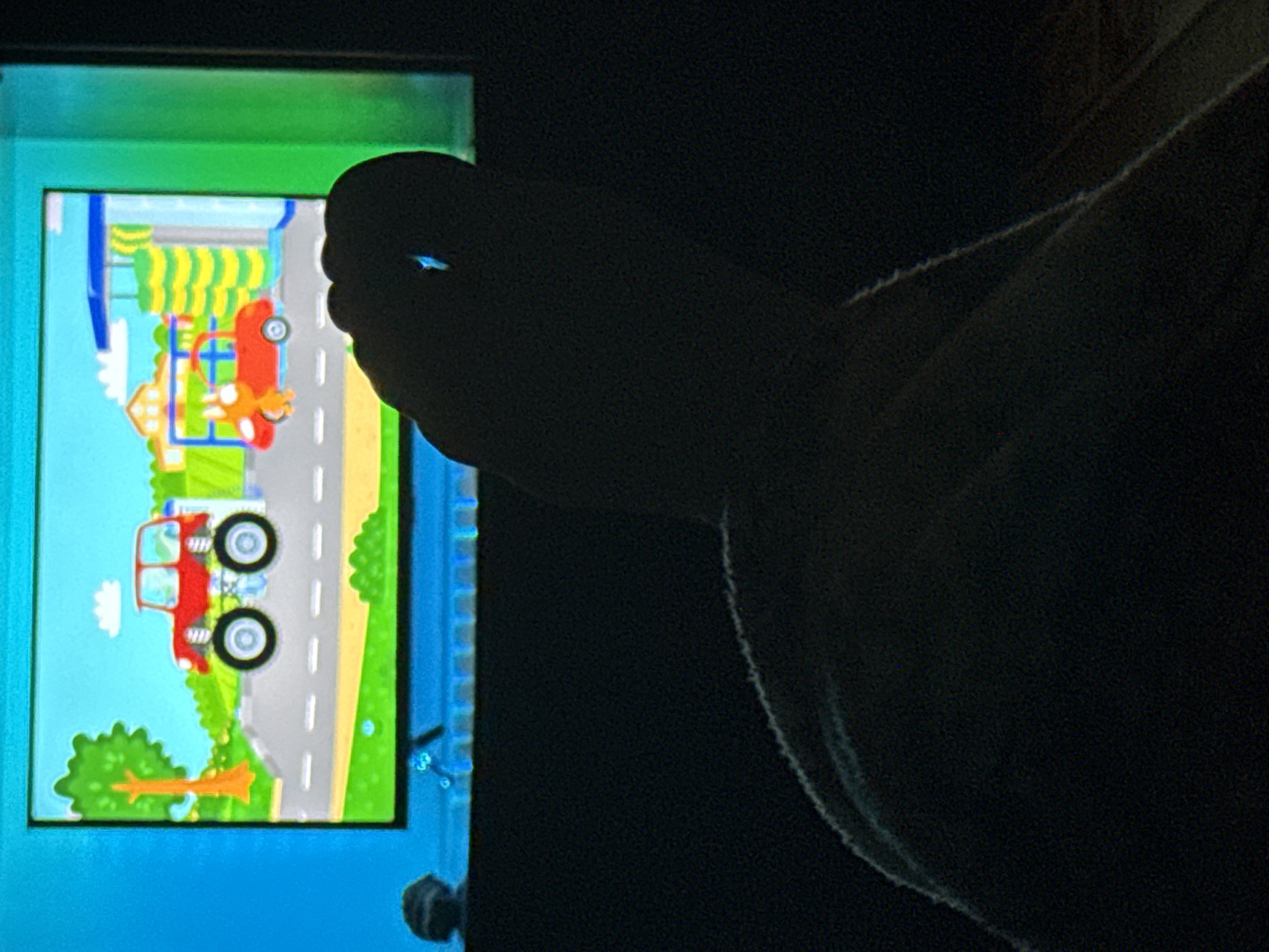 In the dimly lit interior of an Unknown location, an illuminated television screen displays a colorful cartoon scene of vehicles. A red monster truck with large wheels is parked on a road, next to a smaller red car at a car wash. To the left of the screen, a solitary object resembling a small, dark rock rests on a surface. In the foreground, a human foot, silhouetted against the television's glow, is prominently positioned, with the big toe appearing to be in focus. The surrounding environment is dark, with hints of a blanket or fabric visible in the lower portion of the frame, suggesting a relaxed setting, possibly a living room.