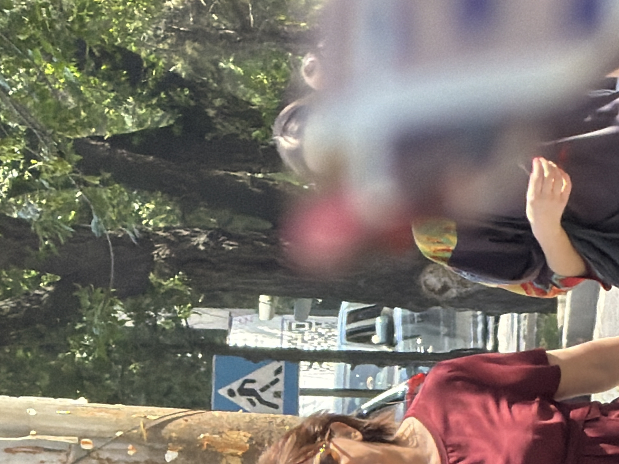 In the city of Unknown, Unknown, a pedestrian crossing sign is visible on the left side of the frame. A woman with brown hair and sunglasses, wearing a red dress, walks on the left side of the road. Behind her, a vehicle is parked. Further in the background, trees and foliage create a shaded environment. A person is visible in the center, wearing a dark garment with a colorful pattern. The scene appears to be outdoors on a sunny day.