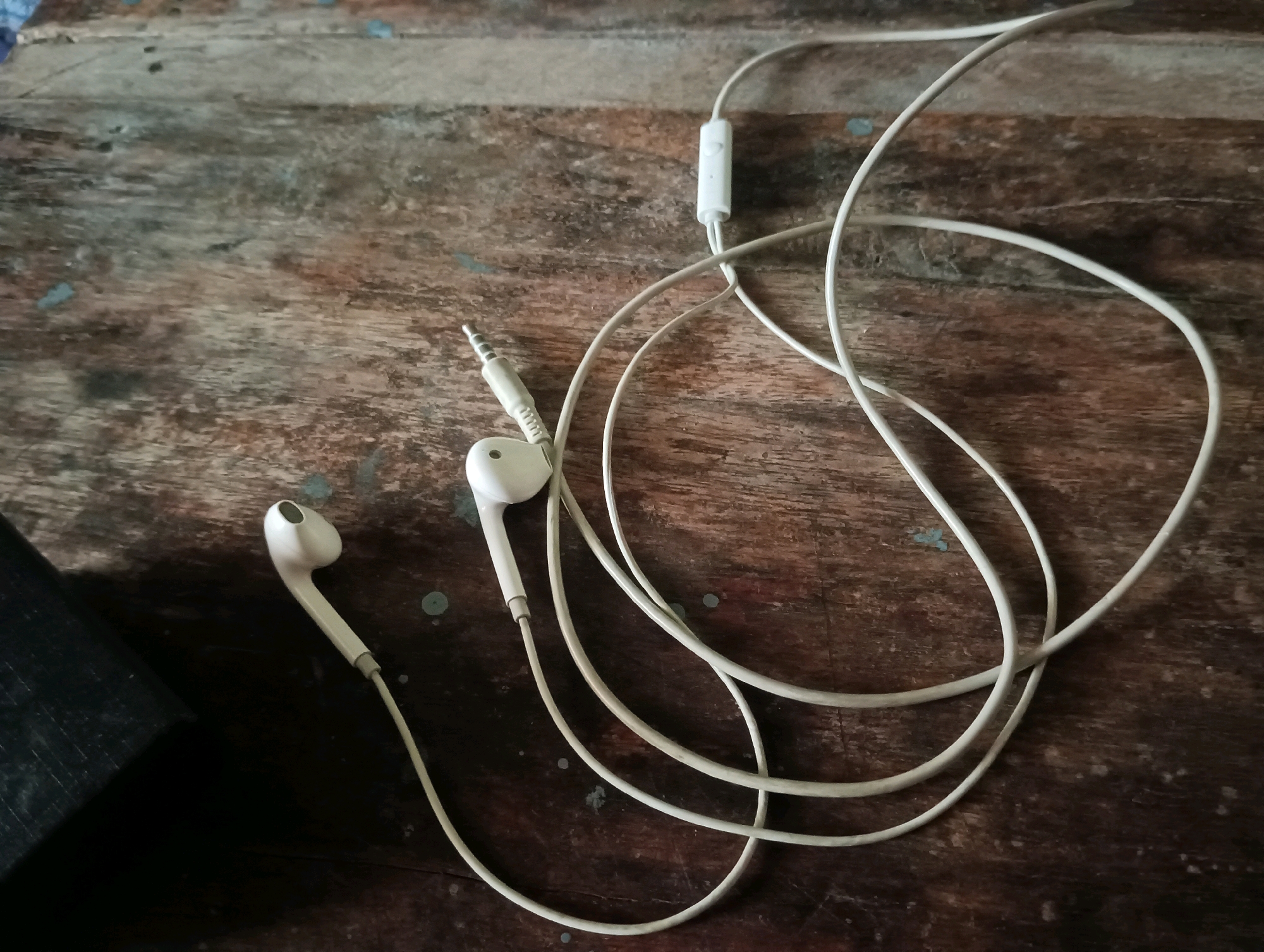 A pair of white wired earphones are laid out on a distressed wooden surface. The earphones are white and connected by a white cord that is partially coiled. A small in-line microphone or control unit is visible on the cord. The wooden surface has a visible grain and appears aged, with some lighter-colored spots and marks on it. A dark, possibly black, rectangular object is partially visible in the lower left corner of the frame. The scene is lit from above and to the right, casting shadows on the wooden surface. The overall impression is of a simple, everyday object placed on a rustic surface. The location is identified as Dakhin Parsa, India.