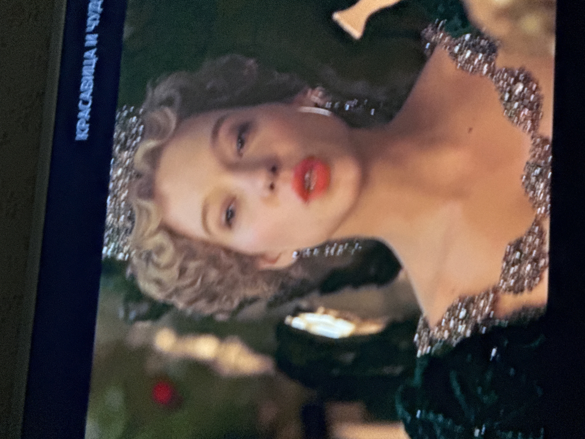 The image shows a television screen displaying a scene from what appears to be a movie or show. The woman on the screen is fair-skinned with curly blonde hair and is wearing a crown and a dark-colored dress with a jeweled neckline. She has red lipstick and is wearing hoop earrings. In the top left of the screen, there's Russian text that translates to "Beauty and the Beast." The environment surrounding the woman on the screen is blurry. The image does not reveal any information about Mytishchi, Russia.