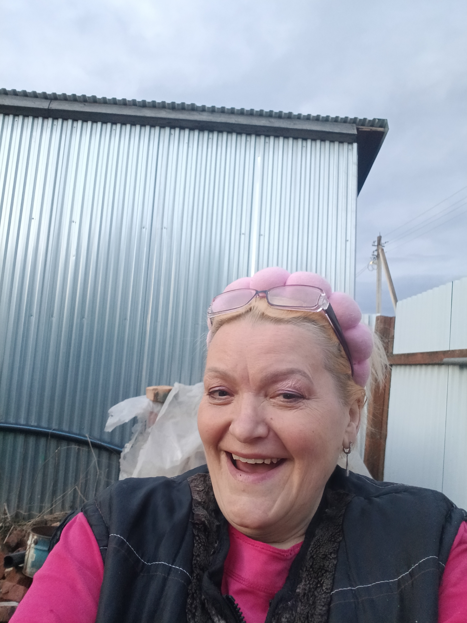 The image features a close-up, eye-level portrait of a cheerful woman positioned outdoors in Красный Маяк, Russia. She has blonde hair and is wearing a vibrant pink long-sleeved top underneath a dark sleeveless vest with a fluffy, possibly faux fur, lining around the collar. A distinctive pink headband, adorned with several spherical elements, sits on her head, with a pair of eyeglasses resting on top of it. She displays a broad, joyful smile, revealing her teeth, and her eyes convey a sense of happiness and amusement, suggesting she is looking directly at the camera, likely for a selfie.
The background consists of a tall, corrugated galvanized metal wall on the left side of the frame, showing vertical grooves and a subtle sheen. To the woman's right, a wooden fence segment is visible, extending towards a utility pole with power lines against a cloudy, overcast sky. In the lower left corner of the background, there is a pile of debris, including what appears to be bricks, a blue bucket, and crumpled white plastic sheeting. The overall setting points to an outdoor, possibly industrial-edge or rural location under an overcast sky, suggesting a daytime scene. No visible text is present in the image.