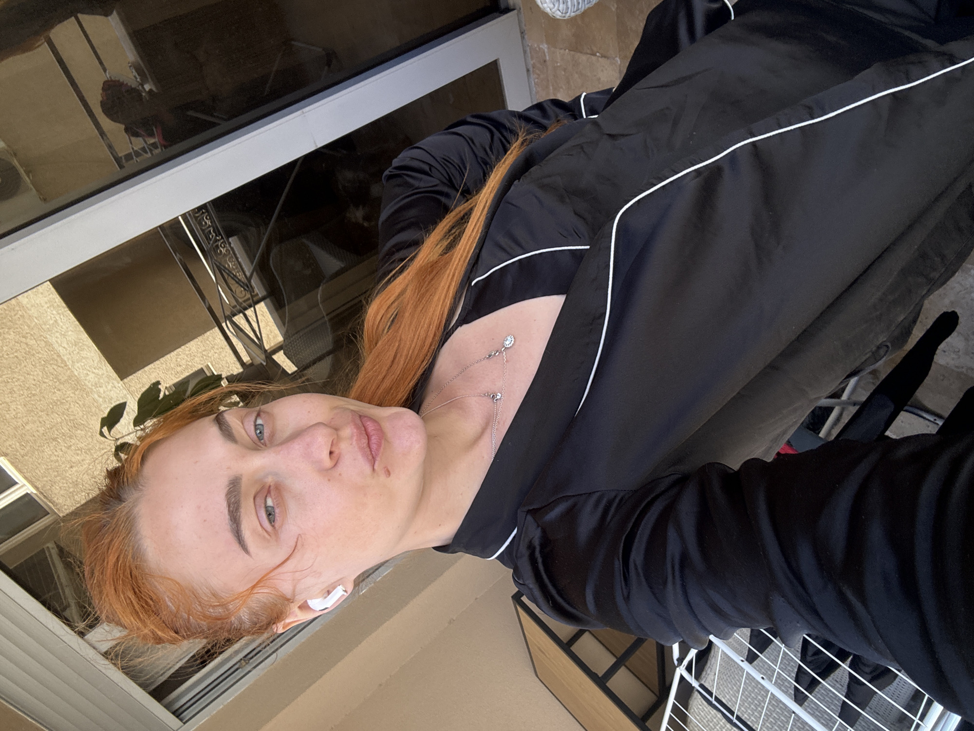 A woman with red hair is posing for a selfie outdoors. She is wearing a black tracksuit with white piping. She is also wearing a necklace and has a white earbud in her left ear. The background appears to be an outdoor patio or balcony with a sliding glass door and a metal drying rack. While the exact location in Budva, Montenegro cannot be determined from the image, the surroundings suggest an apartment or residential area.