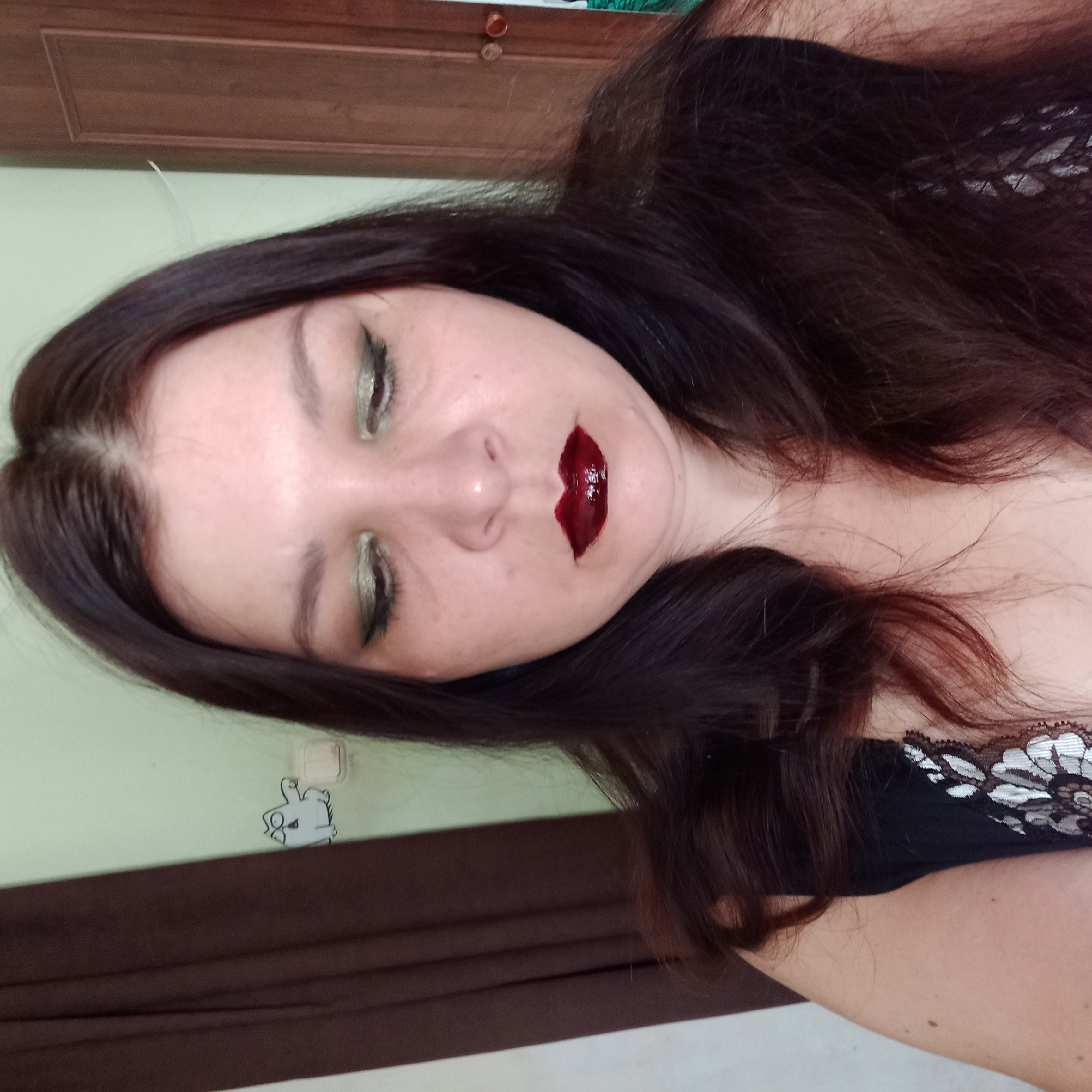 The image shows a close-up of a woman with dark brown hair, wearing dark clothing with white lace detailing. She has dark, glossy lipstick and green eyeshadow. The background is out of focus, but appears to be an indoor setting with a light green wall and a brown curtain. A light switch and a cartoon sticker of a cat are visible on the wall behind her. The lighting suggests an artificial light source. The image does not provide any information about the location in the city of Klyntsi, Ukraine.