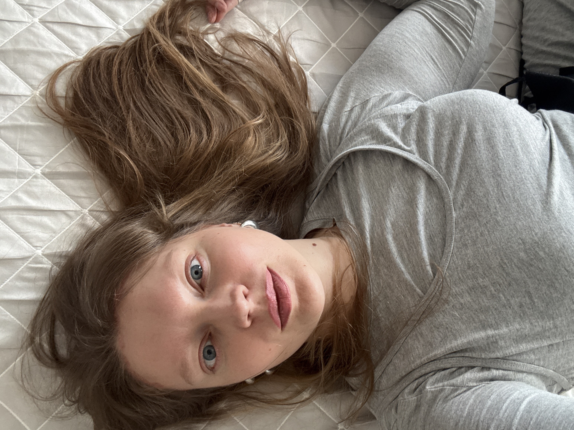 A young woman with light brown hair and blue eyes lies on her back on a white quilted surface, likely a bed. She is wearing a gray long-sleeved shirt. Her hair is spread out around her head. The image appears to be a selfie taken from a high angle. The location is not discernible from the image itself.