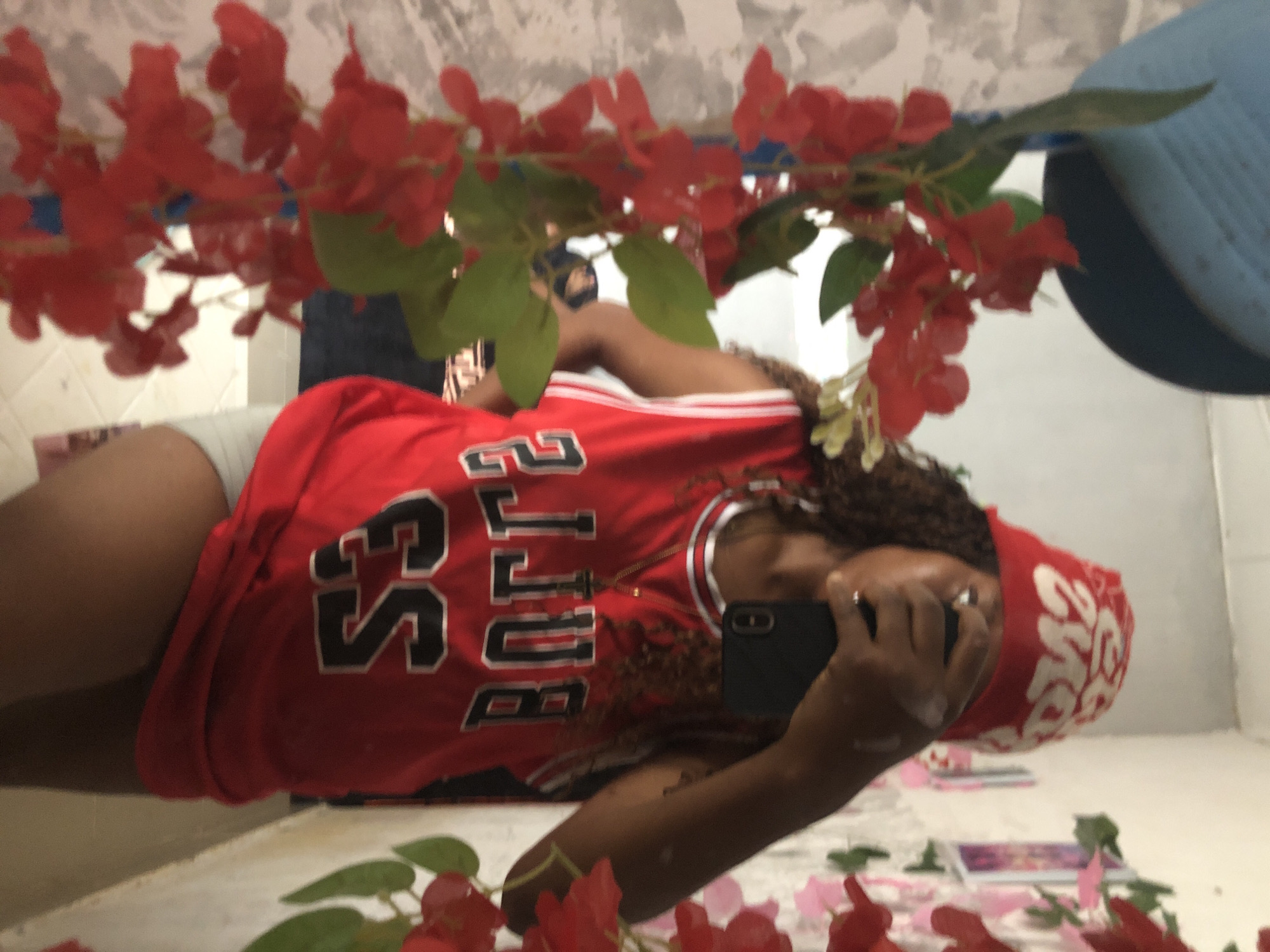 A young woman in a red Chicago Bulls jersey is reflected in a mirror, taking a picture of herself with a black smartphone. She has brown curly hair and is wearing a red bandana with white text on her head. She is wearing gray shorts and has what appears to be a gold chain around her neck. The mirror is decorated with red artificial flowers and green leaves. A blue hat is visible in the upper right corner of the mirror. There are more red artificial flowers and green leaves lying on a white surface below the mirror, along with a few small books and pink petals. The background is a white wall. The photo appears to be taken indoors. The location in the city of Kashere, Nigeria is not evident.