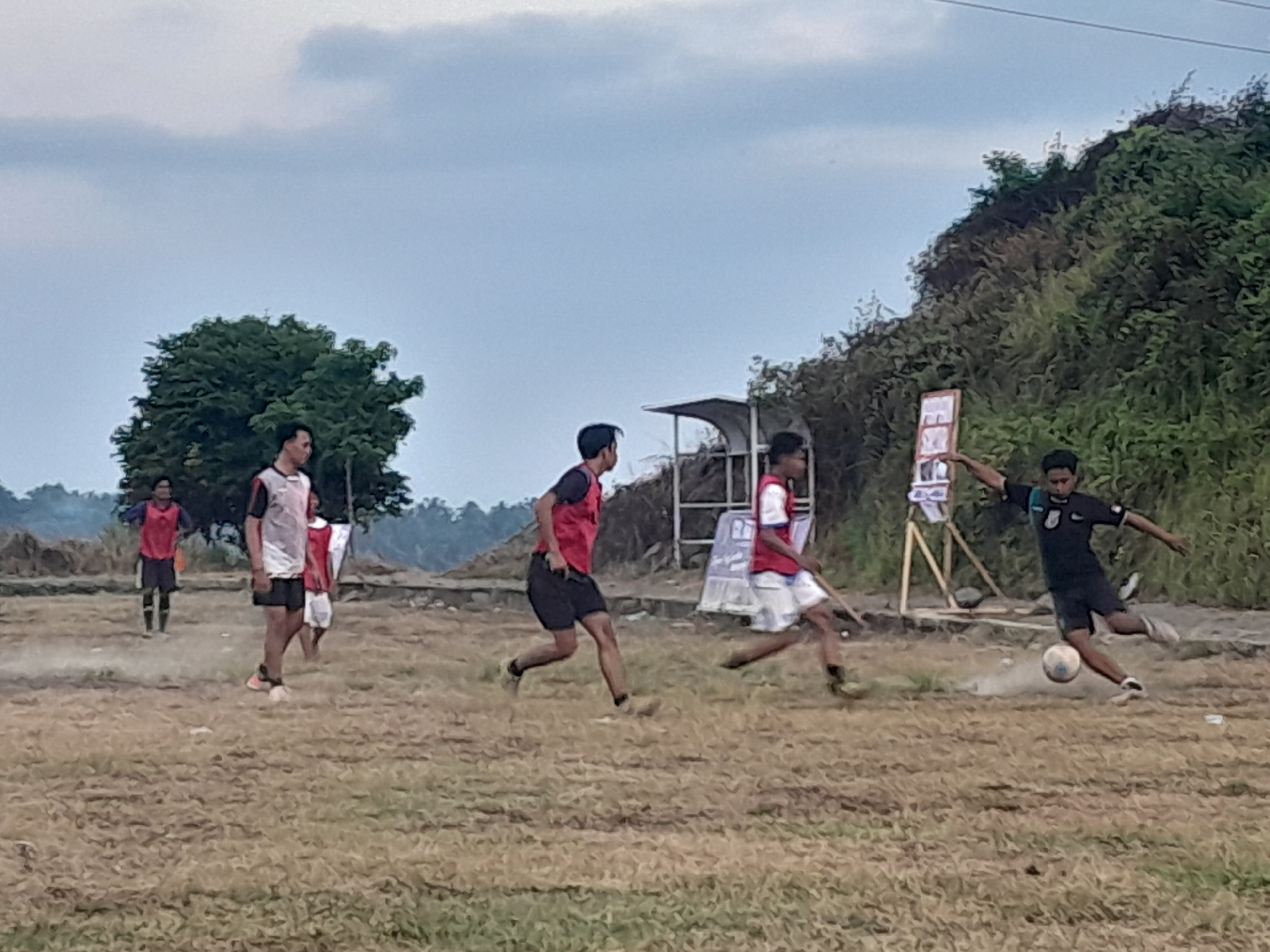 A group of young men are playing soccer on a dry, dusty field in Genteng, Indonesia. The sky is overcast with gray clouds. The field is rough and uneven with patches of dry grass. In the background, there are some trees, a small covered shelter, and a grassy hill covered in dense vegetation.
Several players are visible in action. One player in a black jersey and shorts is kicking a soccer ball, creating a cloud of dust. Another player in a red and black jersey and black shorts is running. A third player, wearing a white and red jersey with black shorts, is also running on the field. In the background, a player in a red jersey and dark shorts stands with his hands on his hips. Another player in a white jersey and shorts is visible further in the background. A sign is propped up on wooden stands behind the players. The overall impression is of an informal soccer match being played in a rural or semi-rural setting.