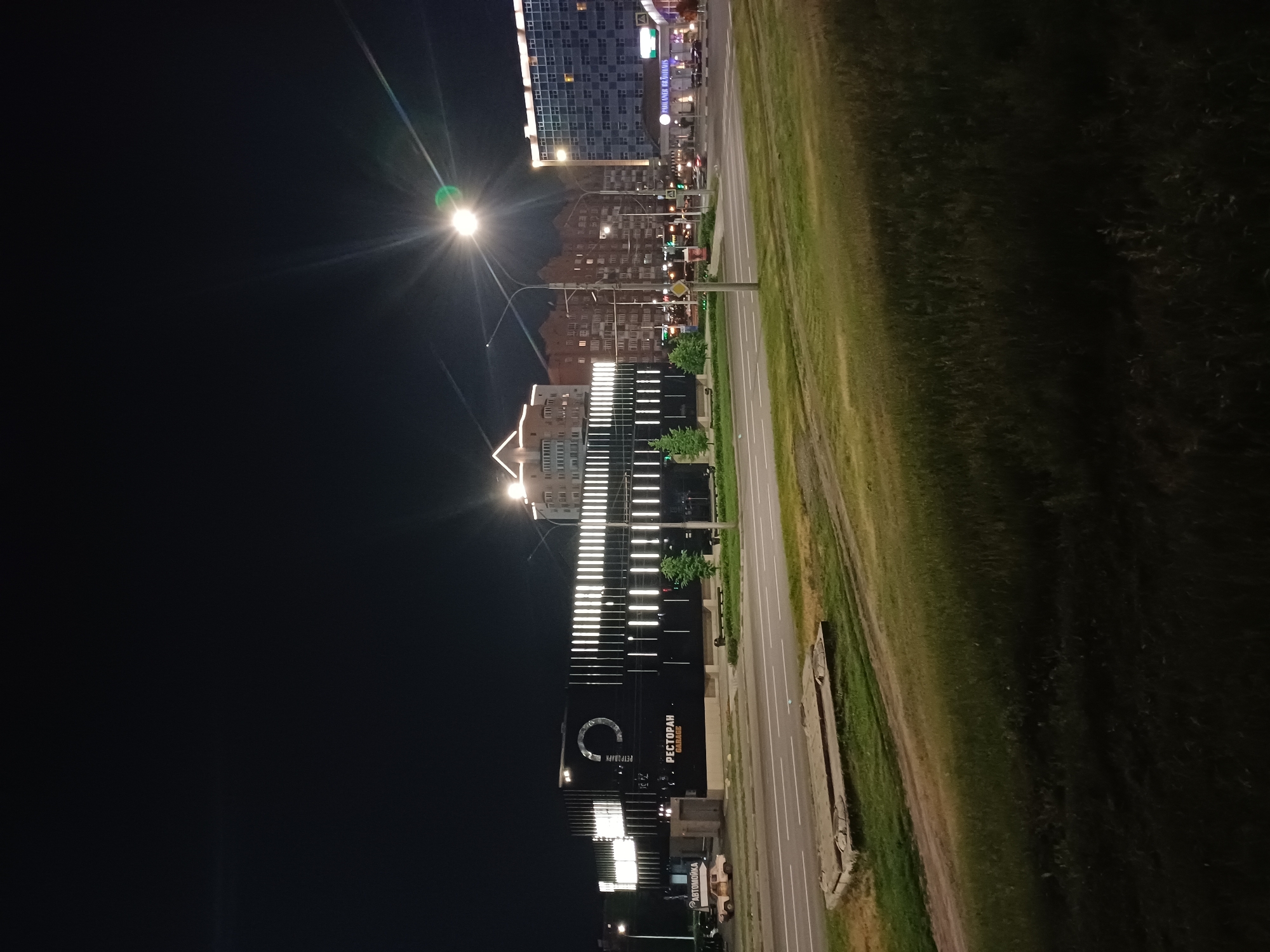In an urban nighttime scene, a wide road with visible lane markings is in the foreground, bordered by a grassy embankment. Across the road, a modern, dark building with a distinctive illuminated facade stands prominently. The building features a large circular logo and vertical light strips, with the word "РЕСТОРАН" (Restaurant) and "GARAGE" visible in illuminated letters. To the left of this building, a smaller structure labeled "АВТОМОЙКА" (Car Wash) is partially visible, with a light-colored car parked in front of it. Further down the road, several other buildings are visible, including a multi-story residential building with many illuminated windows and a taller blue-tinted building on the right. A street light with bright glare is positioned centrally in the frame, illuminating the scene. The overall impression is one of a well-lit commercial and residential area at night.